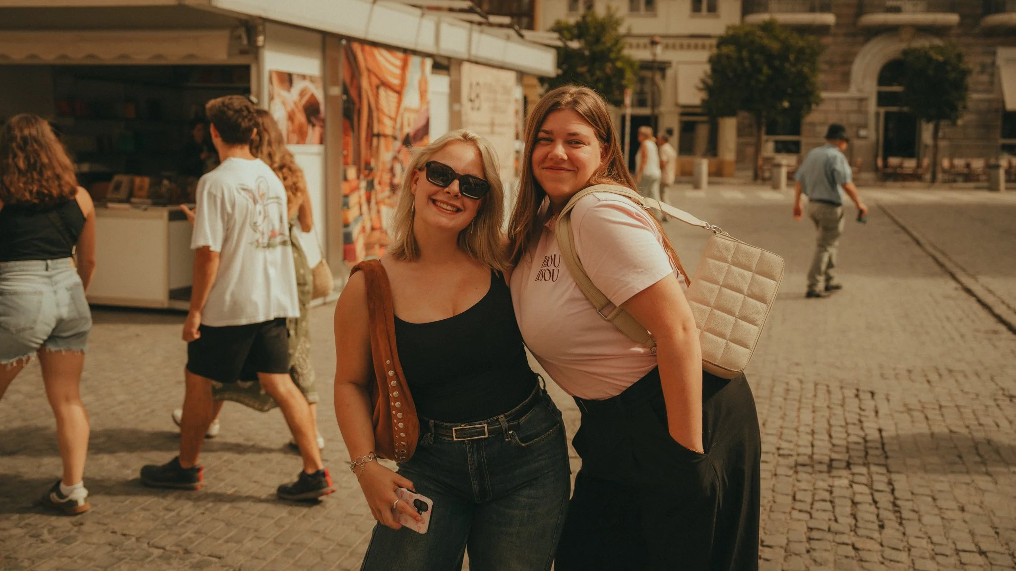 Two women smiling and posing together on a cobblestone street, with others walking and shopping in the background.