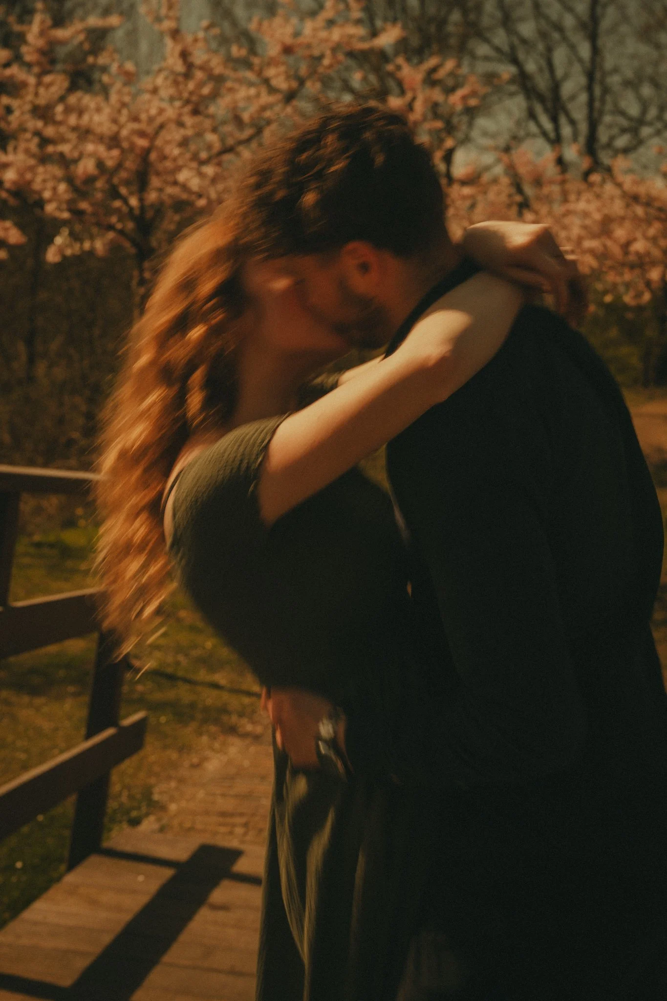 A couple sharing a kiss outdoors during evening or night with pink blossoms and trees in the background, and a wooden pathway in the foreground.