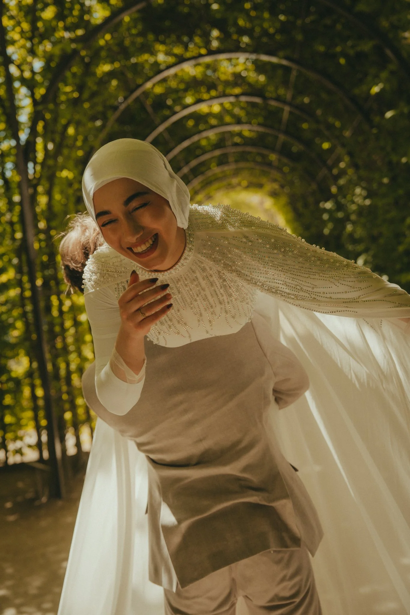A woman dressed in an elegant white gown with detailed beadwork and a matching hijab, laughing and leaning forward with her hand near her face, inside a tunnel of green trees with sunlight filtering through the leaves.