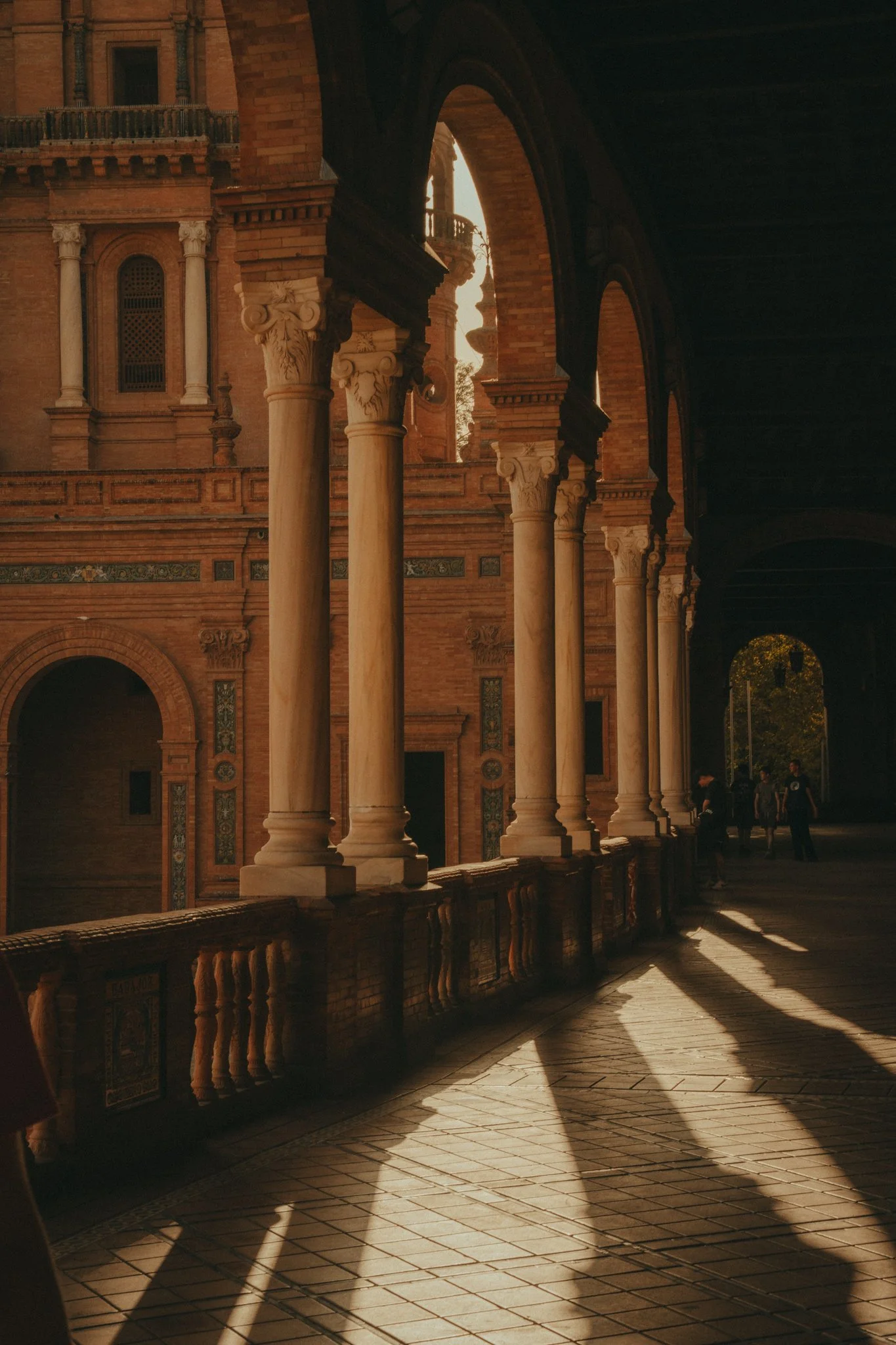 Sunlight casting shadows on the columns and tiled floor of a historic brick building with arches and decorative architecture.