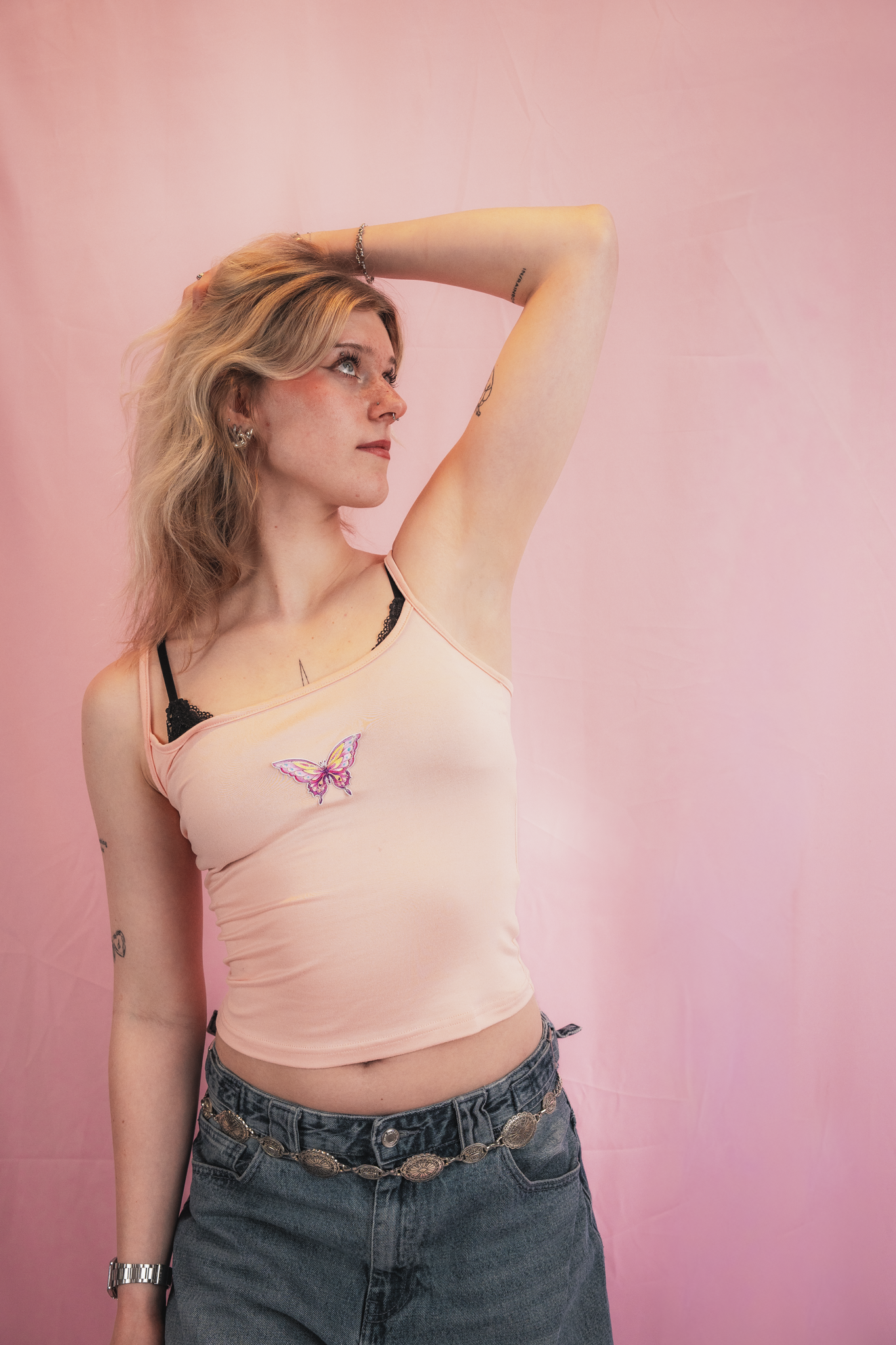Young woman with blonde wavy hair posing against a pink backdrop, wearing a peach tank top with a butterfly design, ripped jeans with a decorative chain belt, earrings, and a watch, with her left arm raised and hand behind her head.