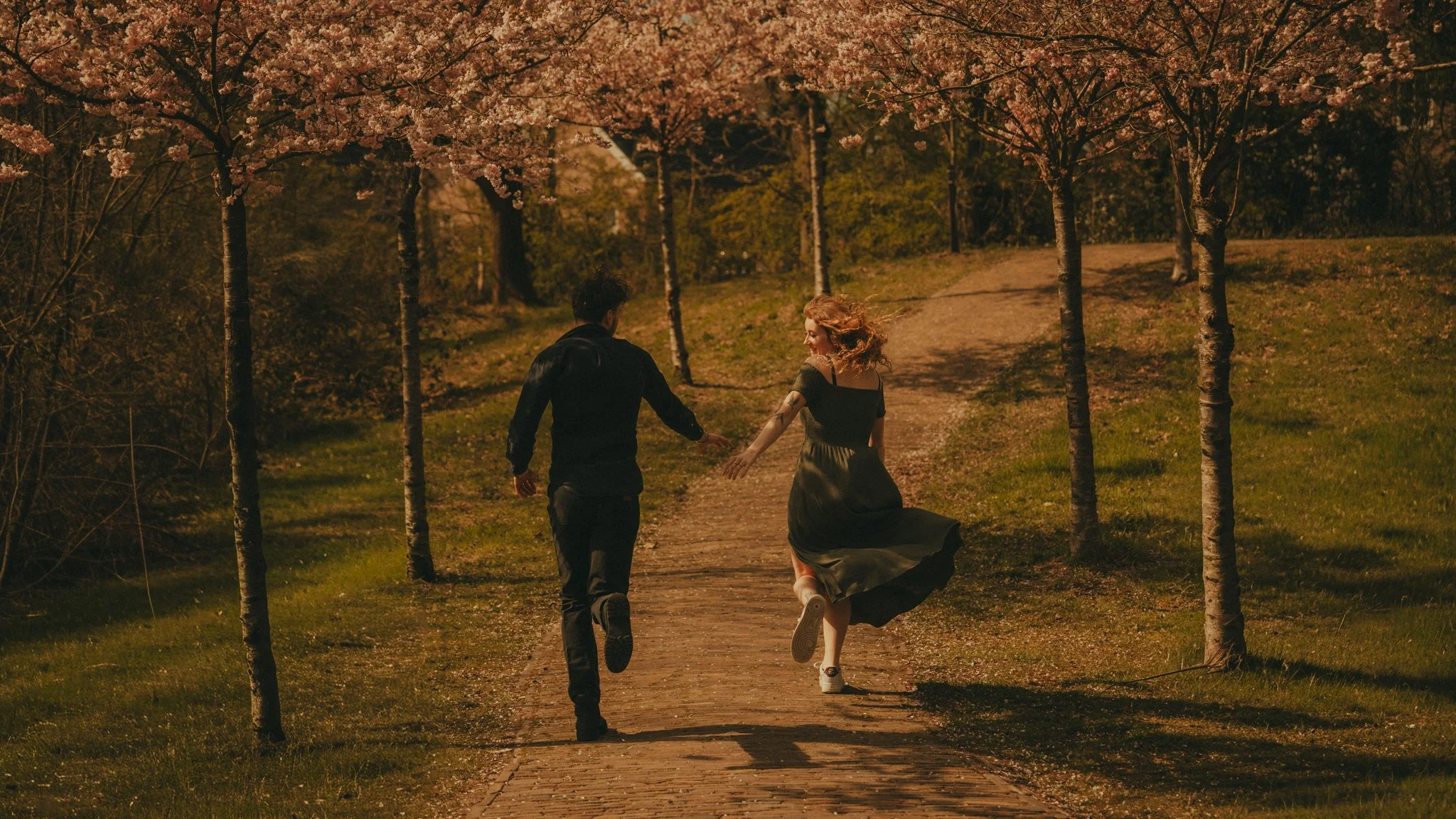 A man and woman holding hands and running through a park with blooming pink cherry blossom trees.