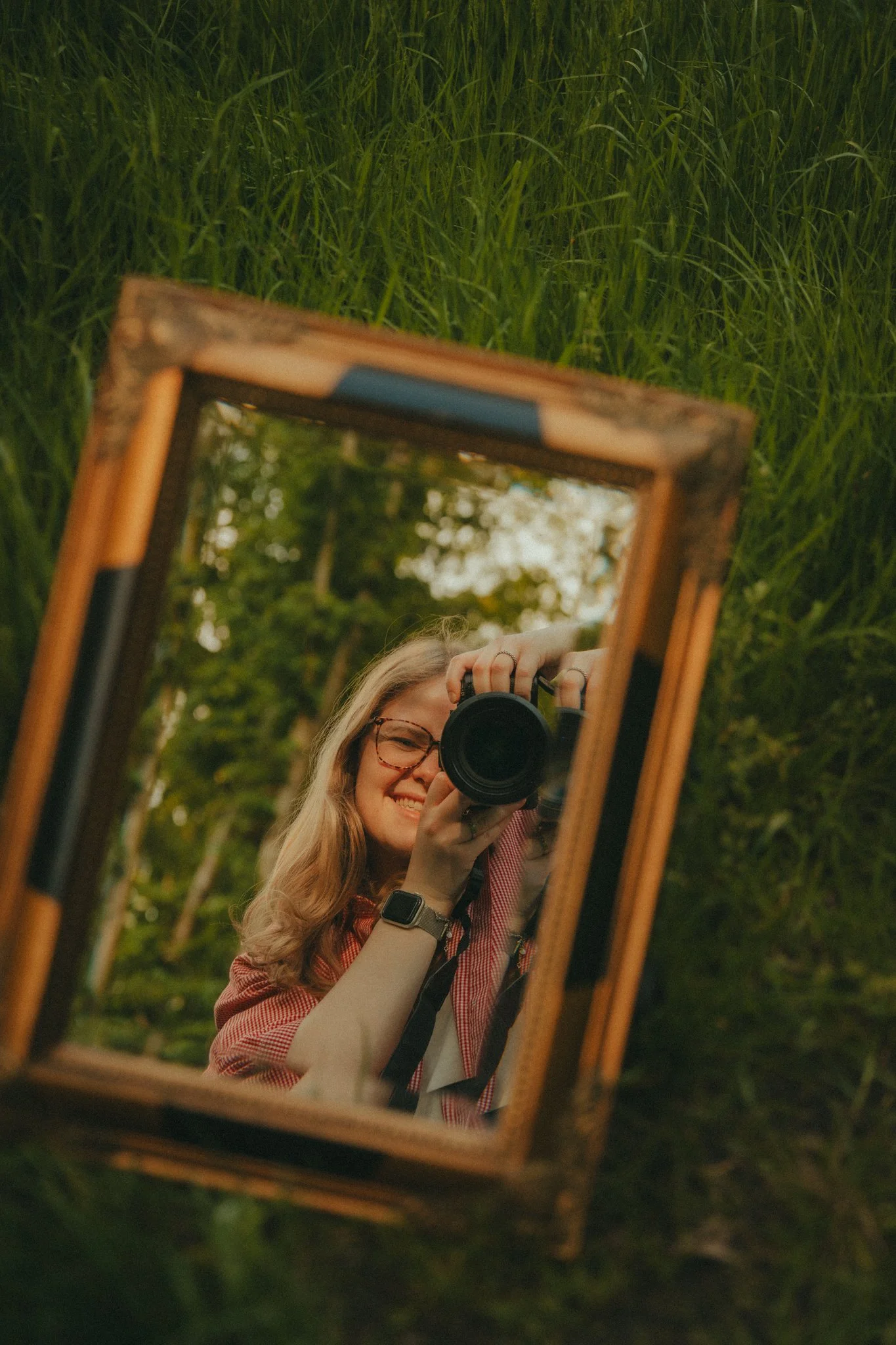 Een vrouw met blond haar en bril neemt een selfie met een camera voor een spiegel die buiten ligt, omgeven door groen gras en bomen.