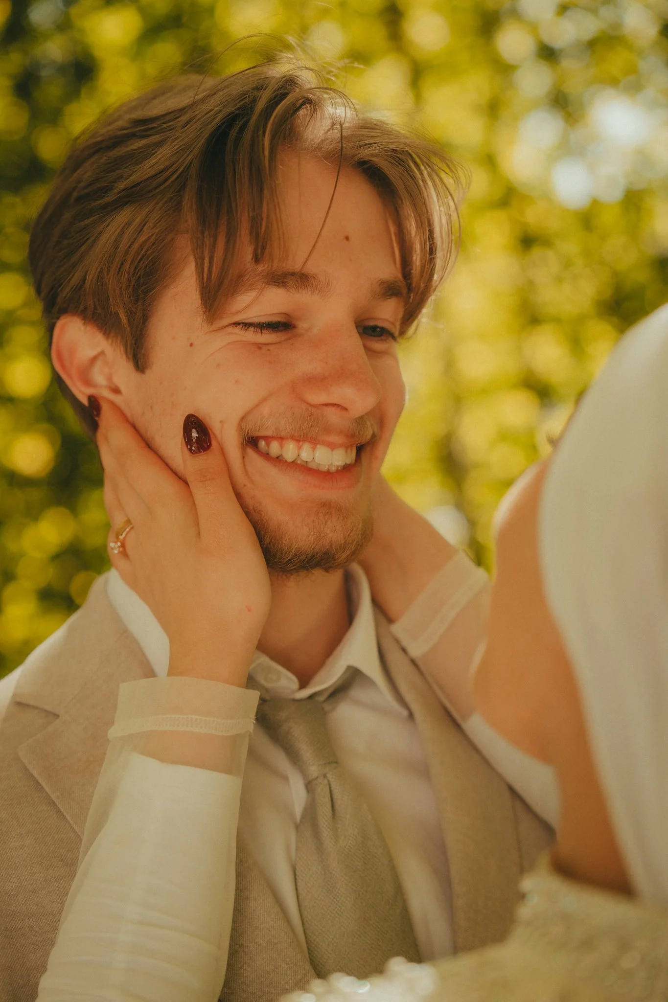 A young man with light brown hair, a beard, and freckles on his face smiling as he looks at a mirror, with a blurred background of yellow and green leaves.