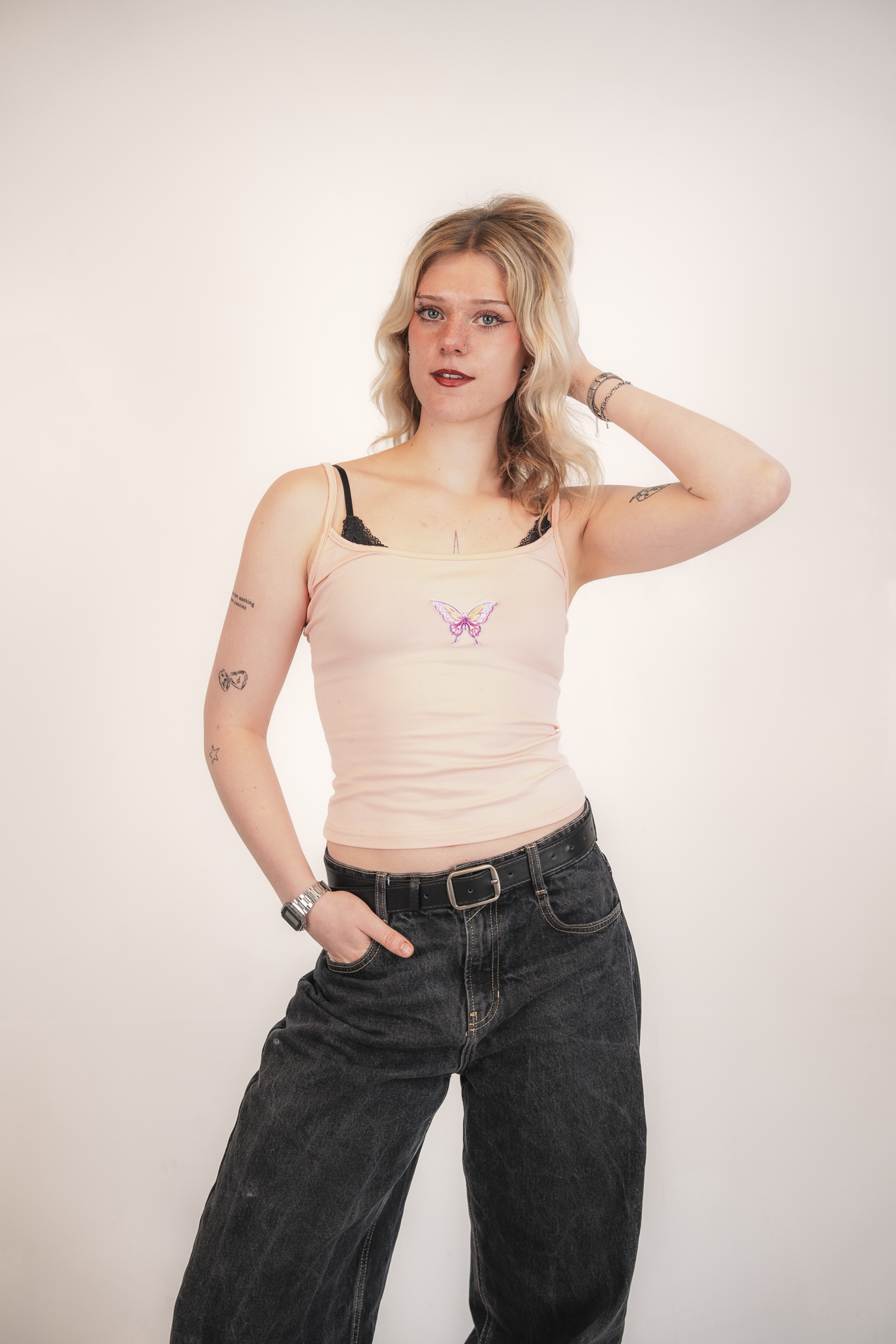 A young woman with blonde wavy hair wearing a light pink tank top with a butterfly design, black lace bralette, black baggy jeans, a silver wristwatch, and bracelets, standing against a plain white wall, posing with her right hand on her hip and her 