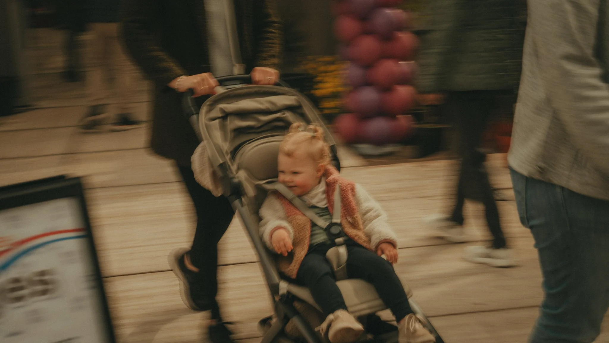 A young girl with blonde hair sitting in a stroller, smiling and looking to the side. The stroller is being pushed by a person whose hands are visible gripping the handle. The background is blurry, suggesting movement, with other people walking by.