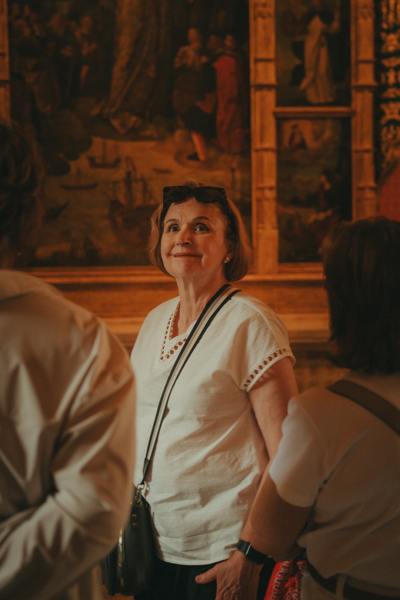 A woman with short brown hair, wearing sunglasses on her head, a white embroidered blouse, and a black crossbody bag, smiling and looking up in a room with historical paintings and wooden walls.