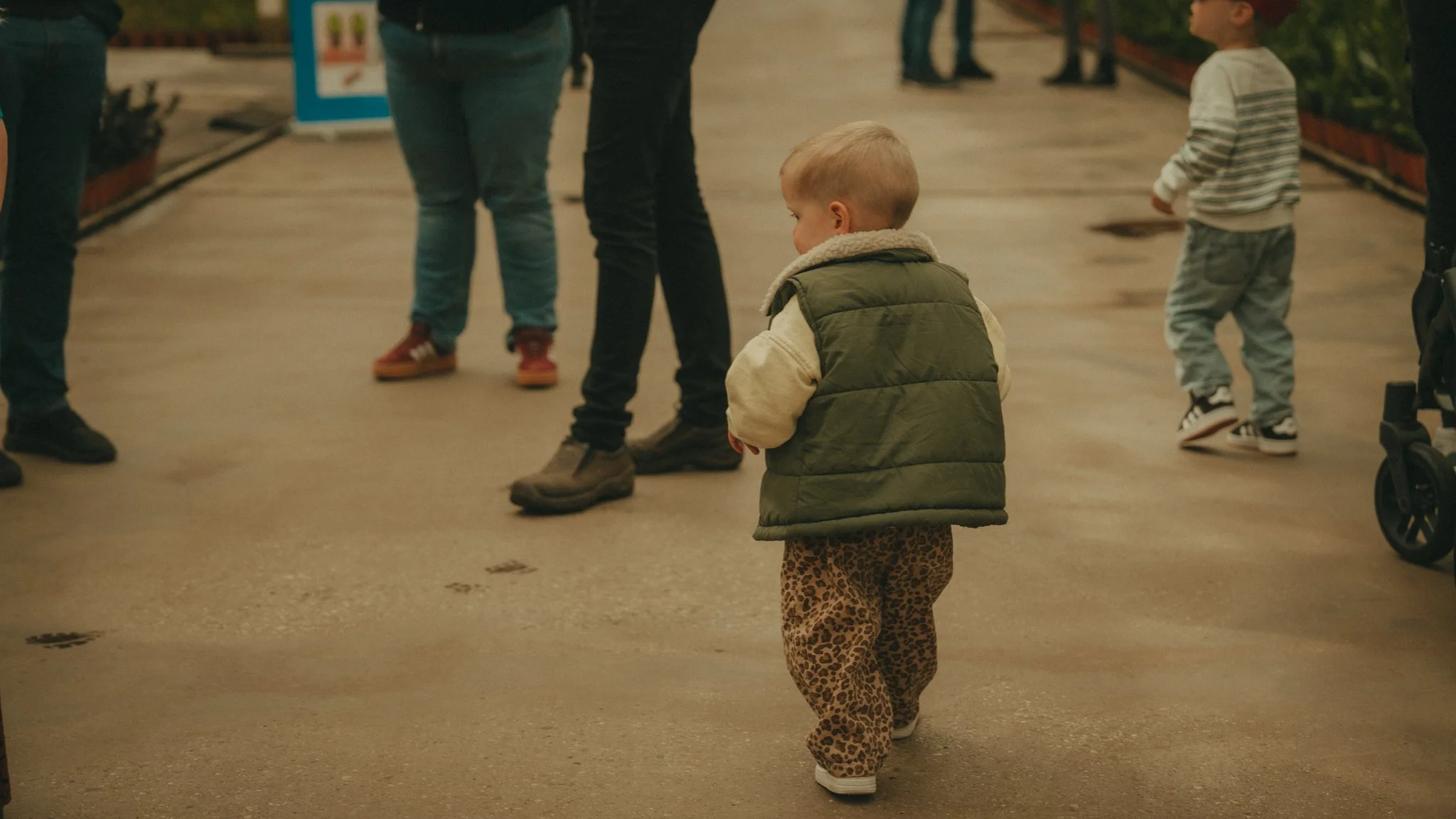 A young child with blond hair wearing a green vest and leopard print pants walking alone in a grocery store aisle, with adults and other children nearby.