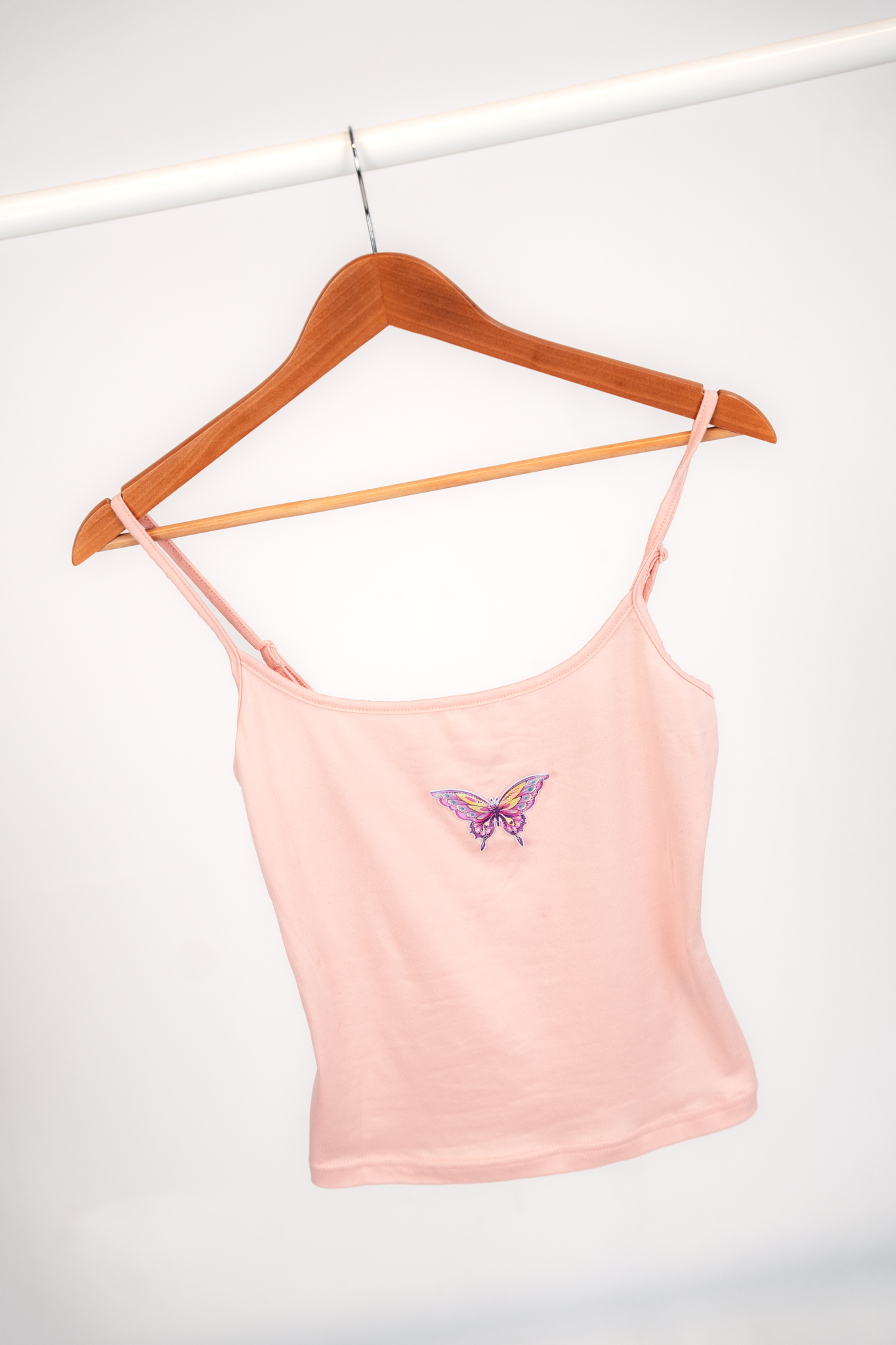 A light pink camisole with a butterfly embroidery hanging on a wooden hanger on a white clothing rail.