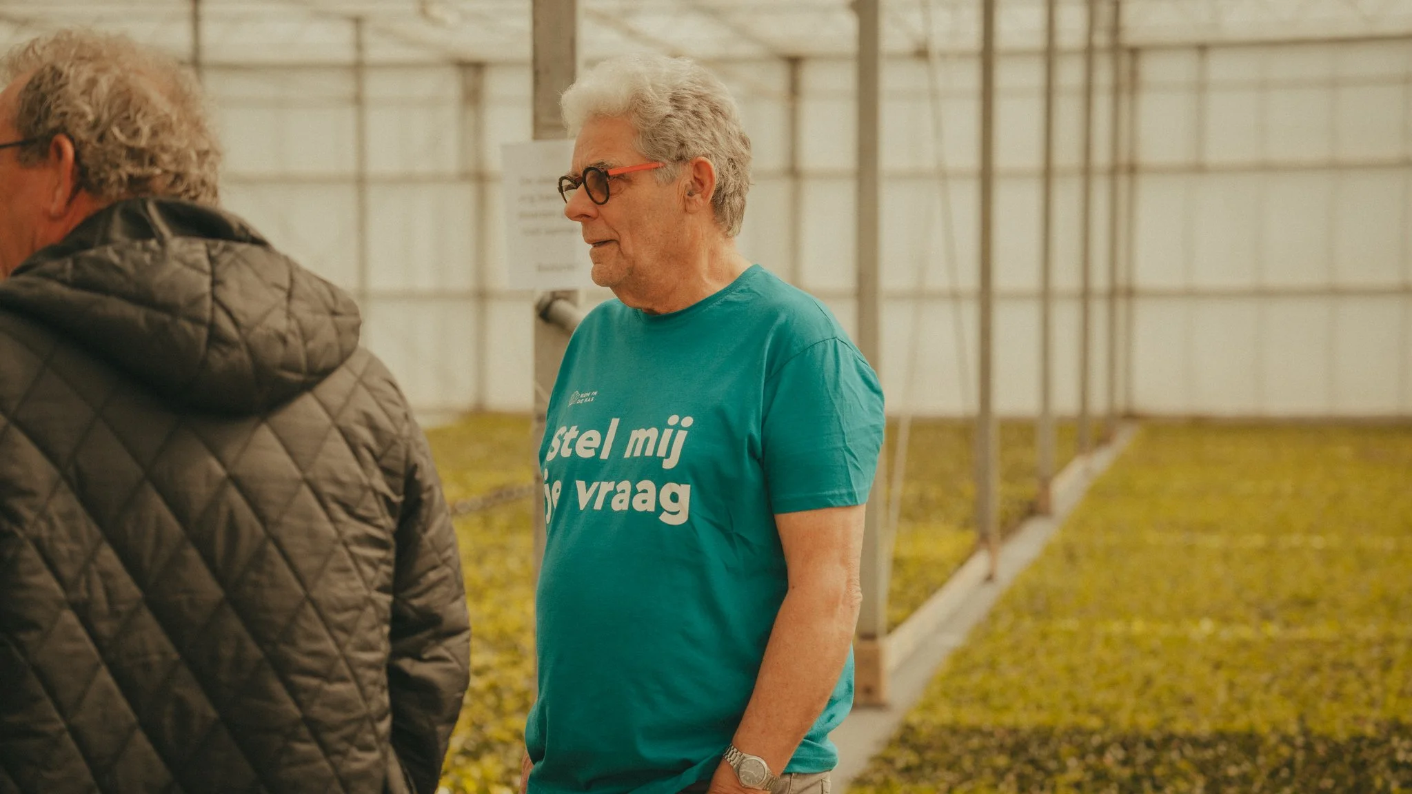 Older man with gray hair, glasses, wearing a teal T-shirt and a watch, talking to another person in a quilted jacket, inside a large greenhouse with leafy plants and a metal framework.