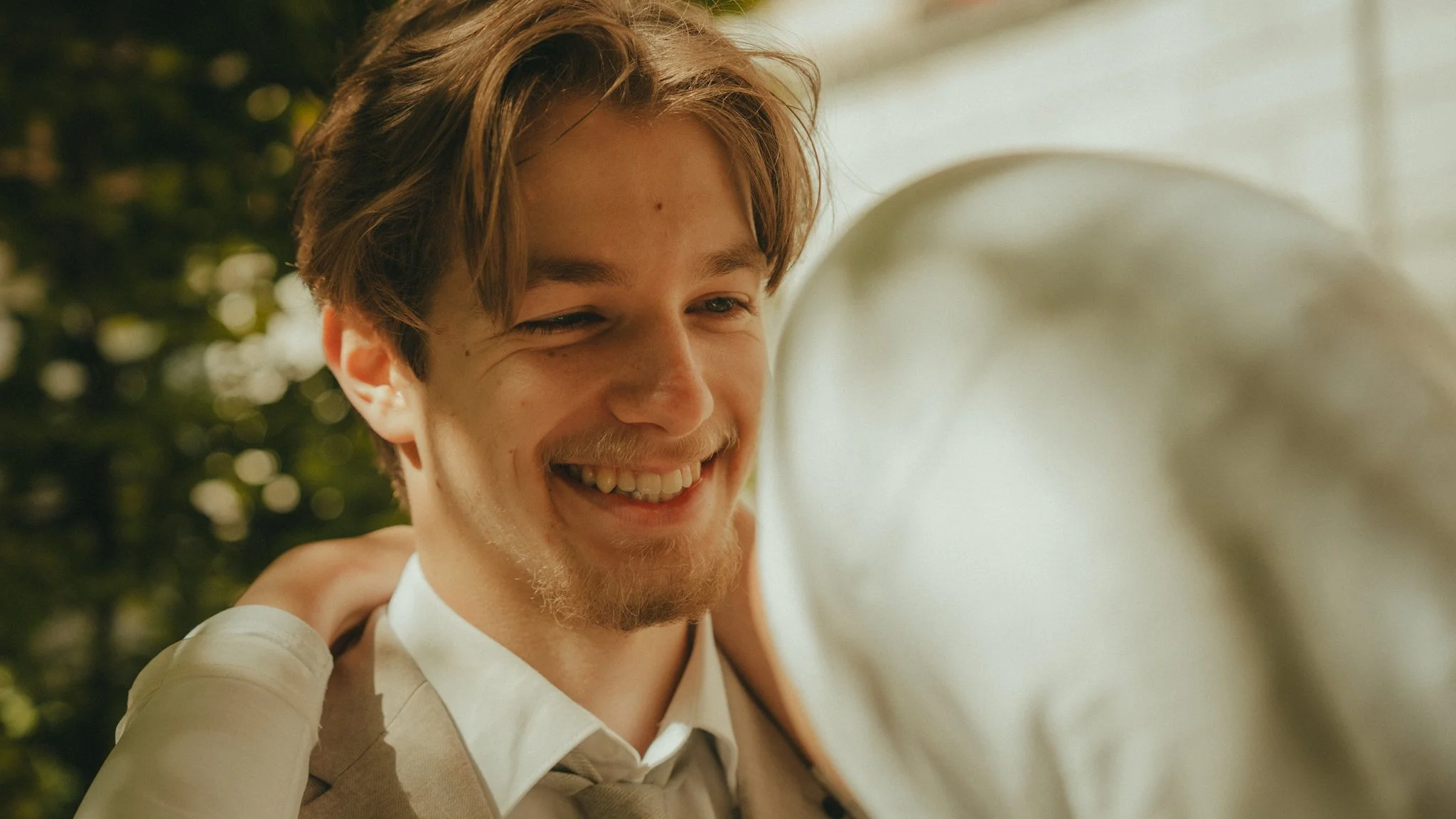 A young man with light brown hair, a beard, and a mustache, smiling with his eyes slightly closed, outdoors in natural light.