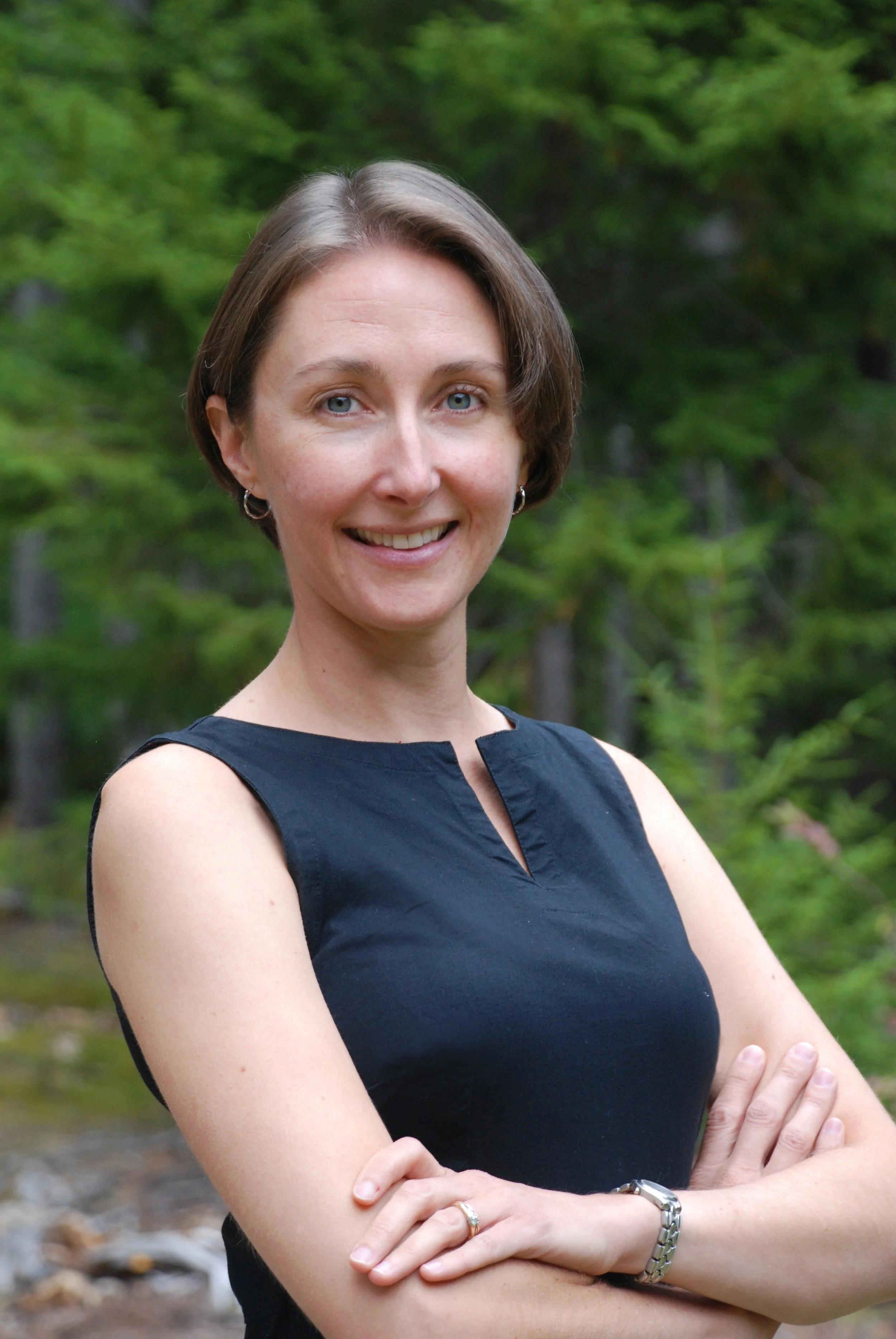 A woman with short brown hair, wearing a sleeveless black top, standing outdoors with green trees in the background, smiling and crossing her arms.