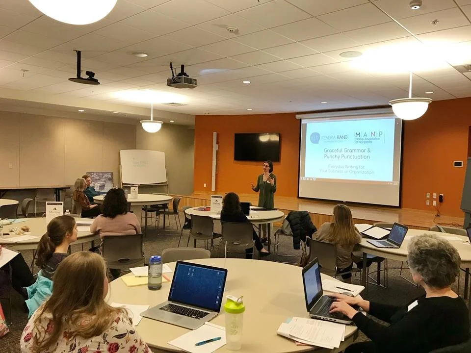 A woman giving a presentation in a conference room with several attendees seated at round tables with laptops and notebooks.