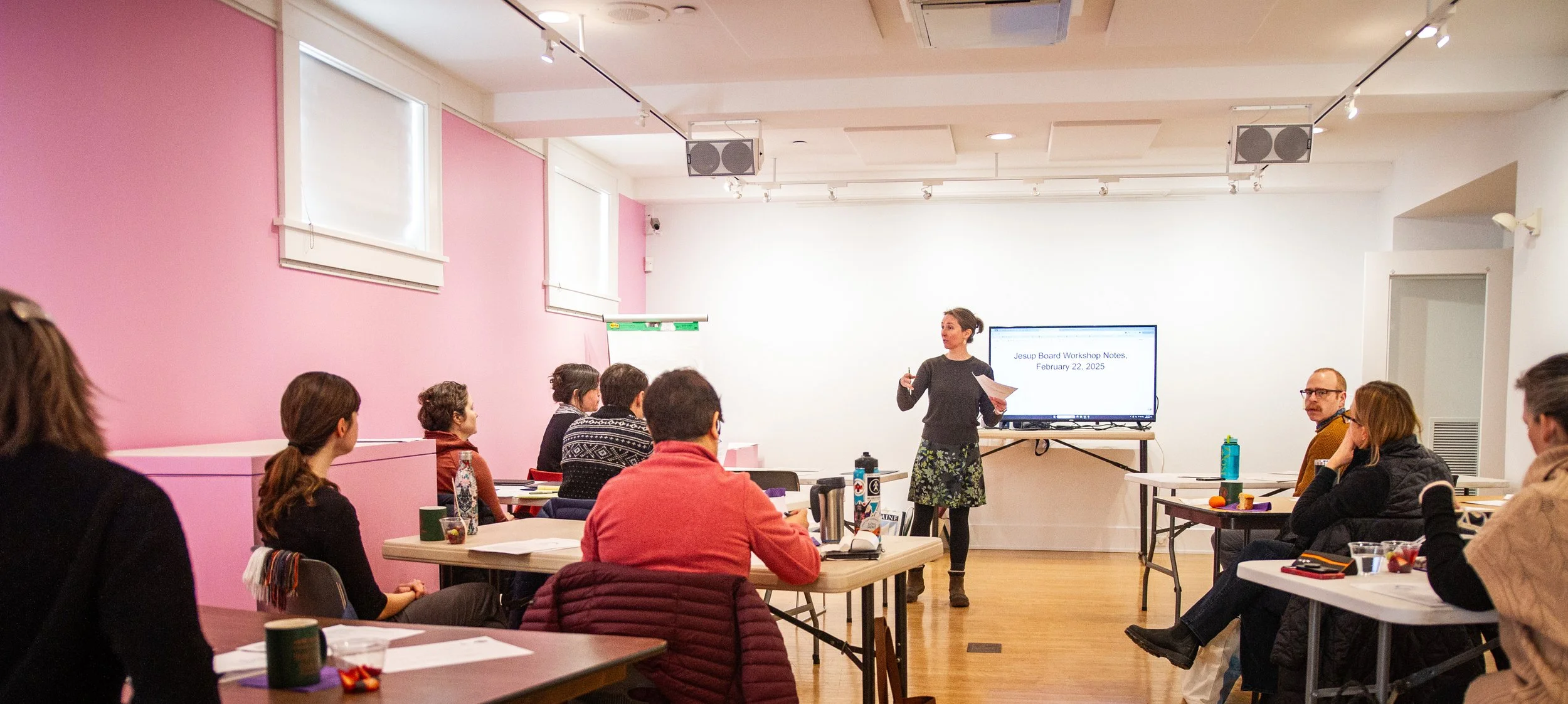 A woman giving a presentation to a group of people in a classroom or workshop setting. The room has pink and white walls, and a large screen displaying notes. The attendees are sitting at tables with various items like notebooks, water bottles, and snacks.