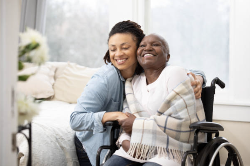 A woman and elderly man in a wheelchair hugging and smiling in a bright room.