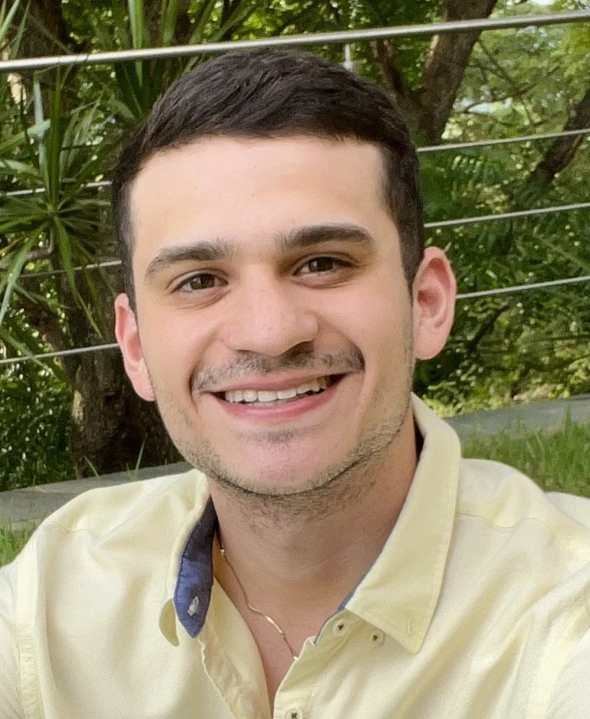 A young man smiling outdoors, with green trees and foliage in the background, wearing a light yellow shirt.