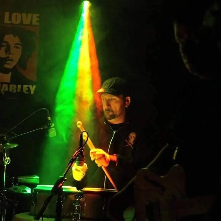 Musician playing drums on stage with colorful rainbow lighting behind him, wearing a black cap and dark clothing.
