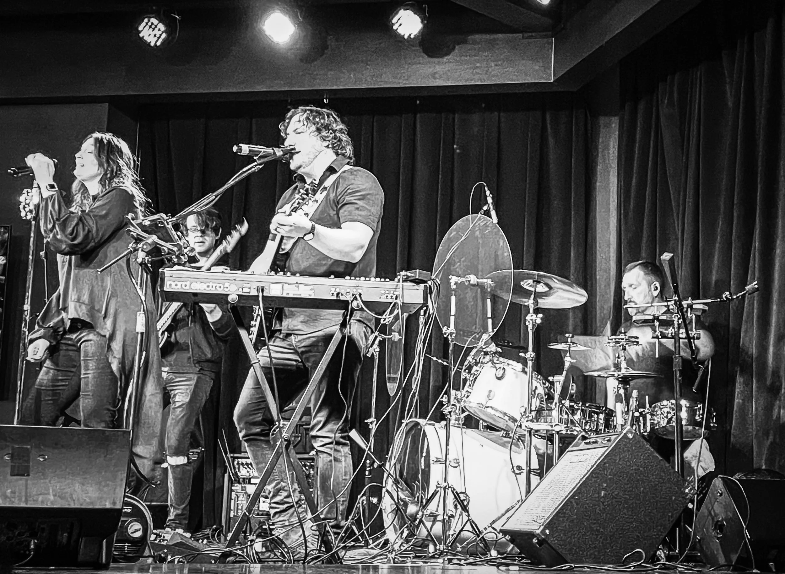 A black and white photo of a band performing on stage, with a singer, a keyboardist, and a drummer surrounded by musical equipment and stage lights.