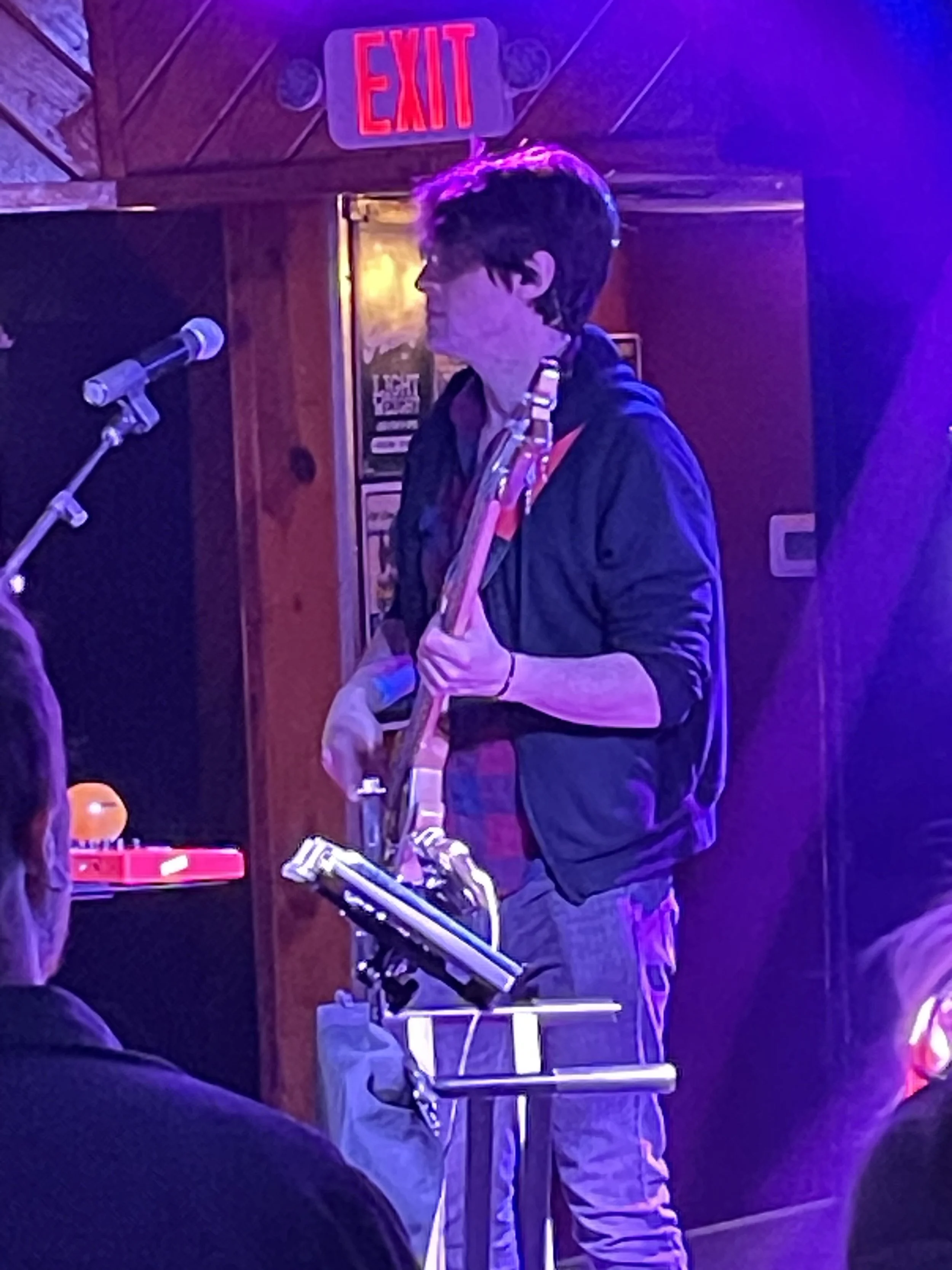 A young musician playing the saxophone on stage in a dimly lit venue with purple lighting, with an audience in the foreground.
