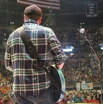 Back of a person wearing a plaid shirt and a cap, with a guitar strapped to their back, at an indoor concert or event.