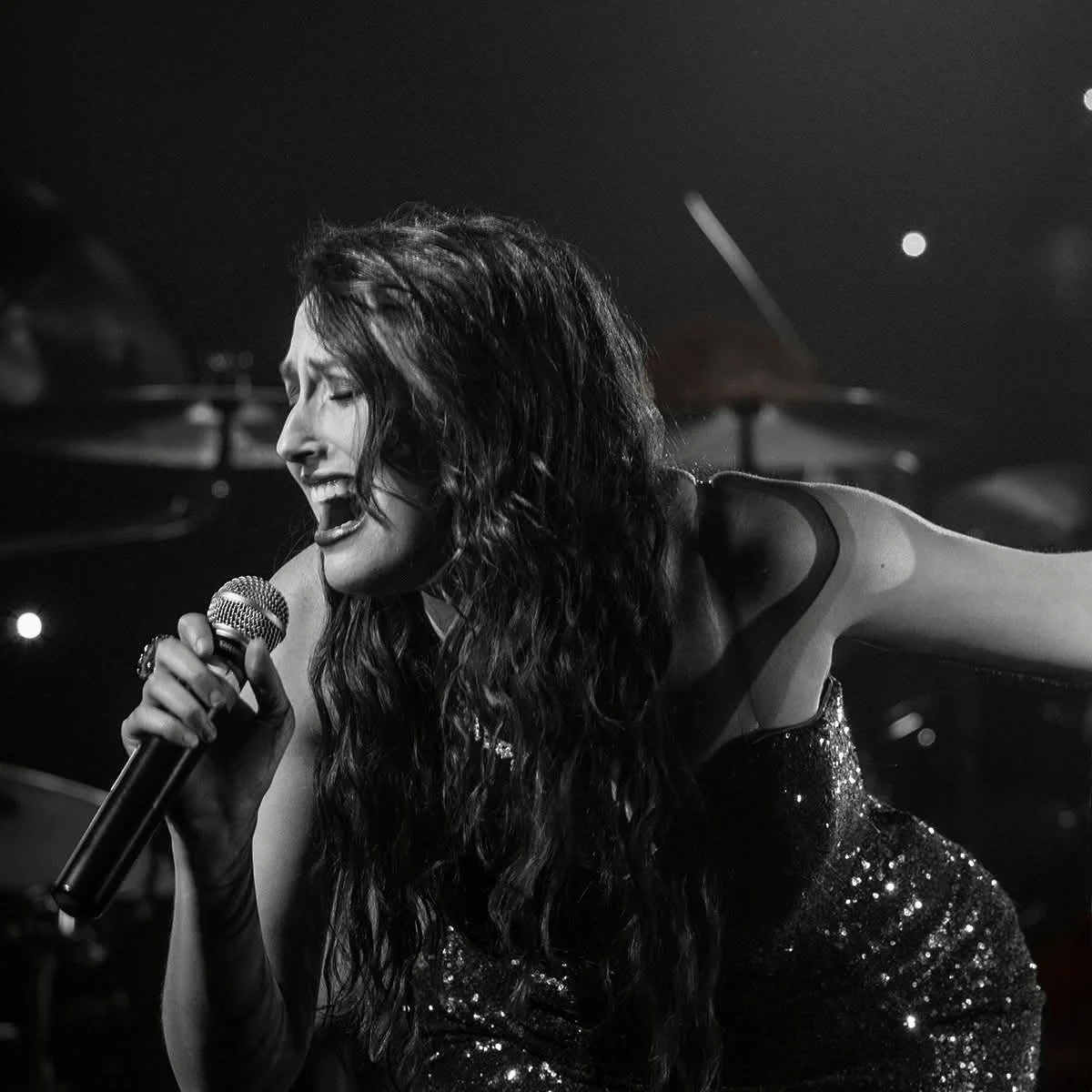 A female singer with long wavy hair passionately singing into a microphone on stage, wearing a sparkly dress.