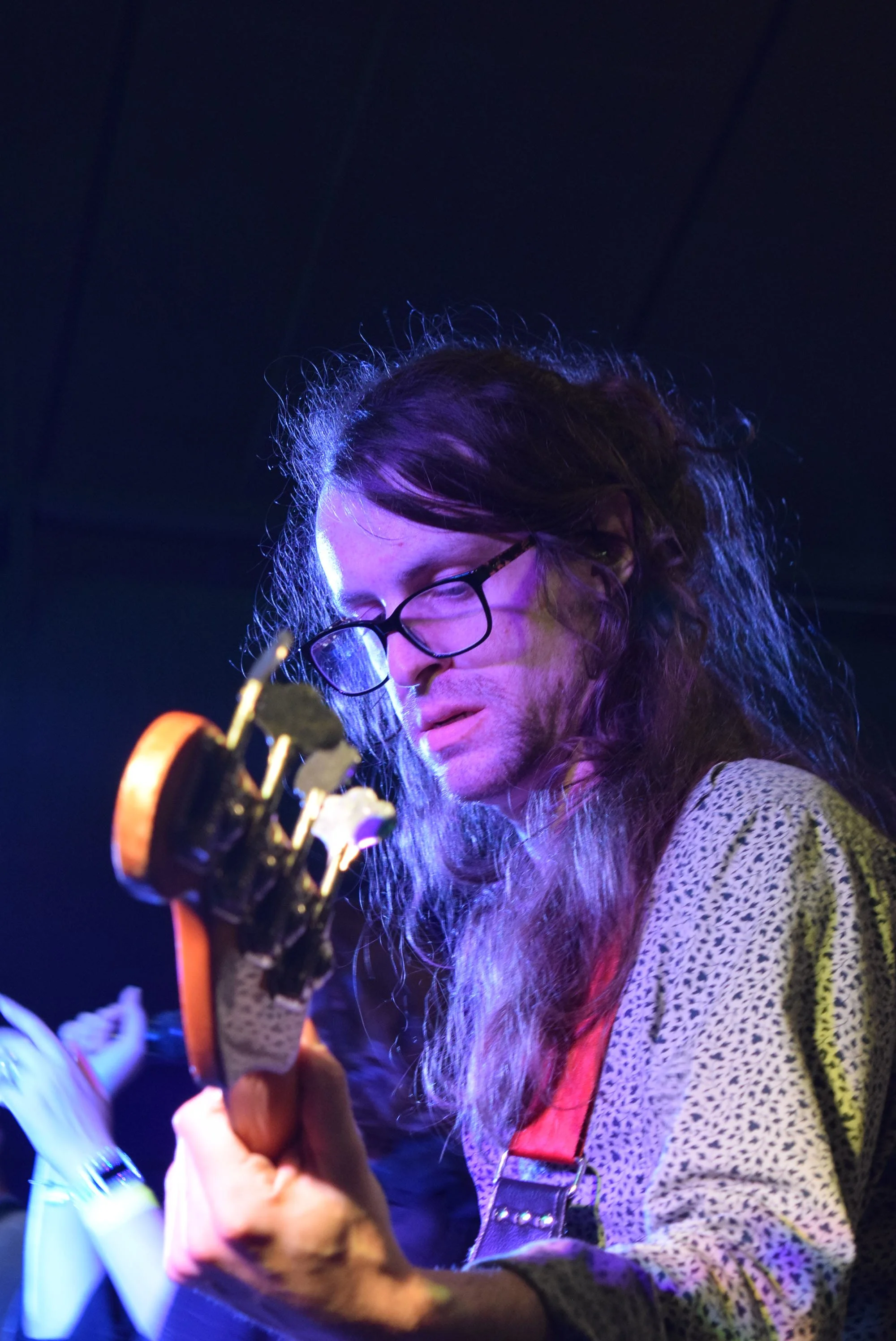 A person with long hair, glasses, and a patterned shirt playing an electric guitar under colorful stage lighting.