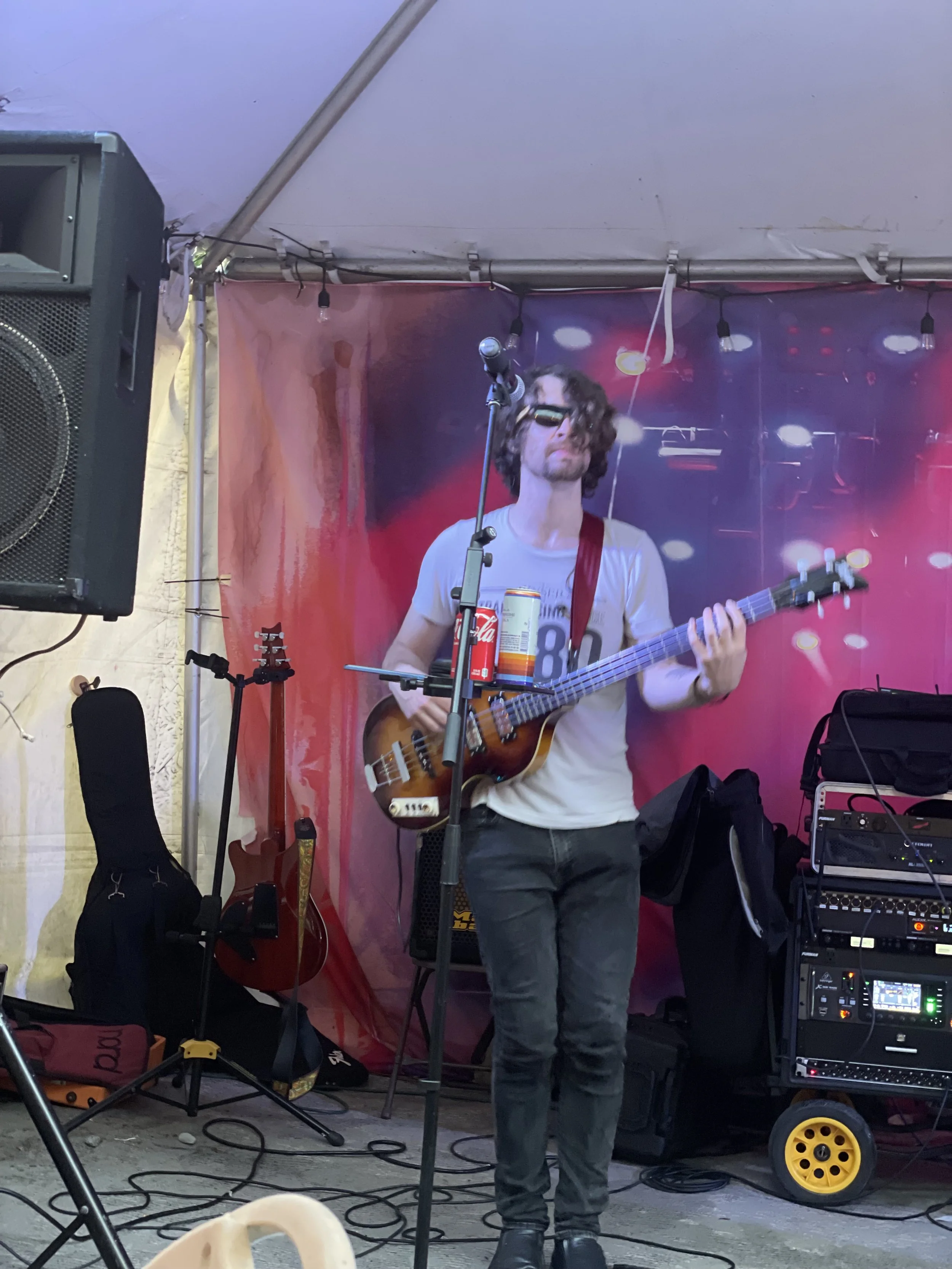 Musician with long curly hair wearing sunglasses, playing an electric guitar on stage with a red strap, surrounded by sound equipment and a colorful backdrop.