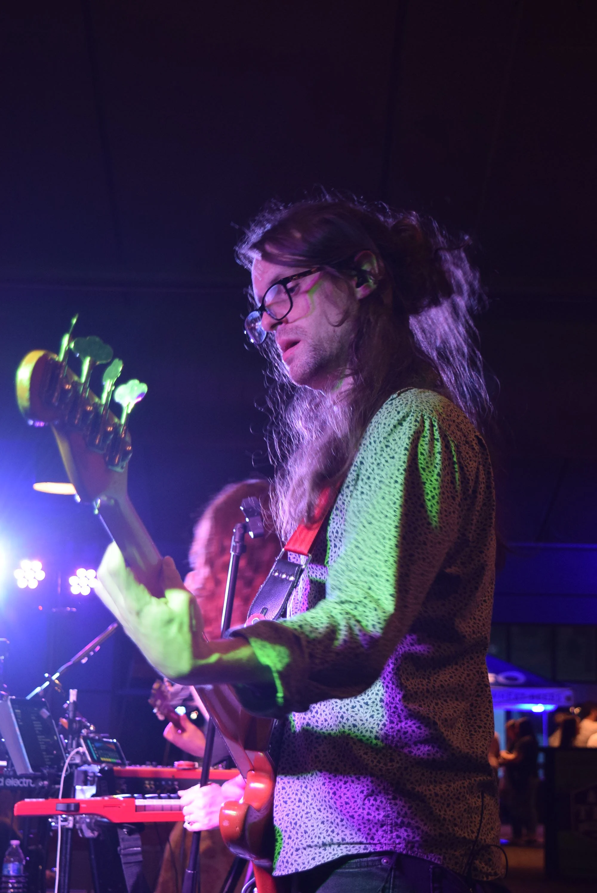 A male musician with long hair, glasses, and a patterned shirt playing an electric guitar on stage with colorful lighting.