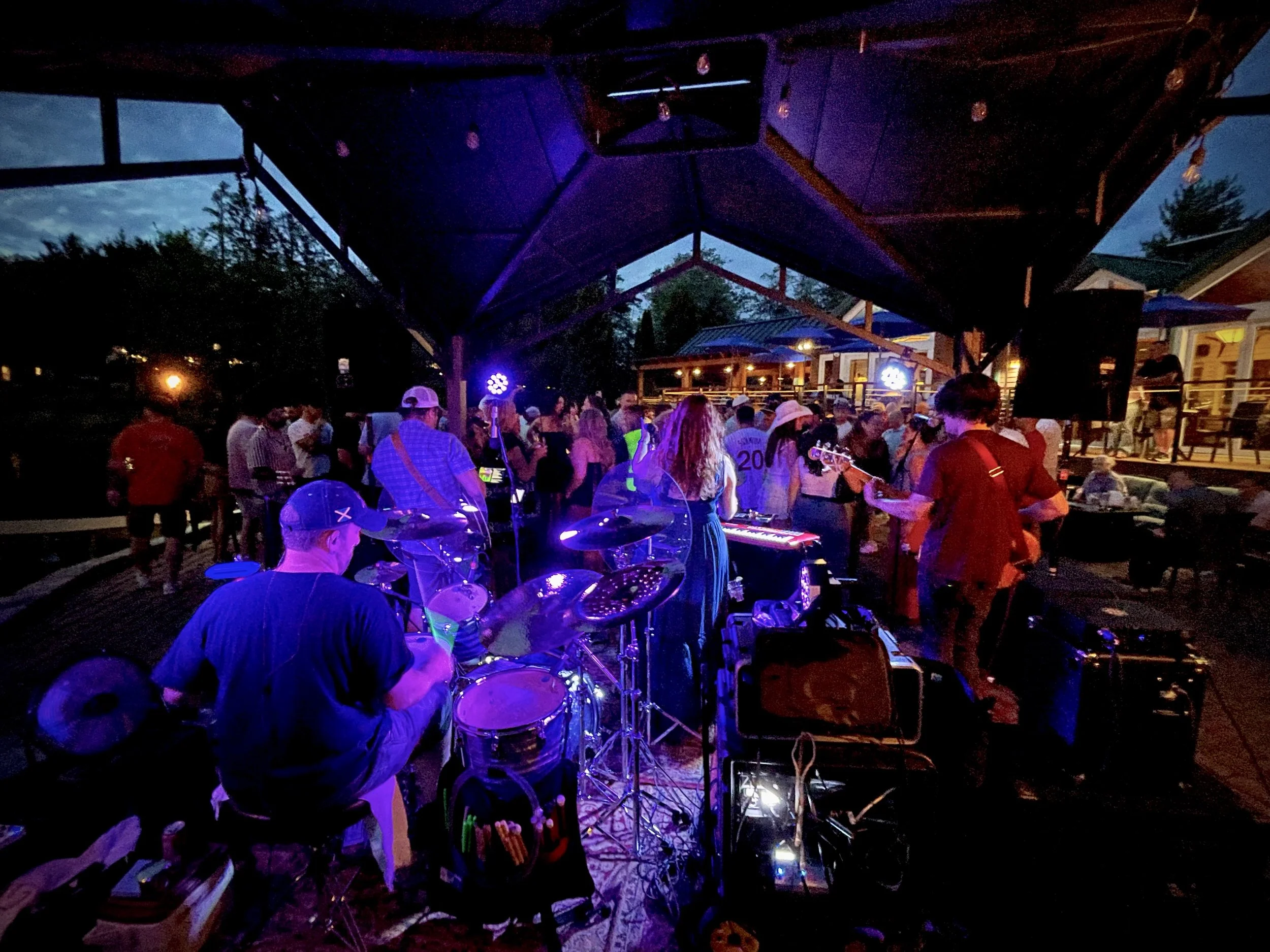 A band playing live music at an outdoor party during the evening, with people dancing and socializing under a covered patio with string lights.
