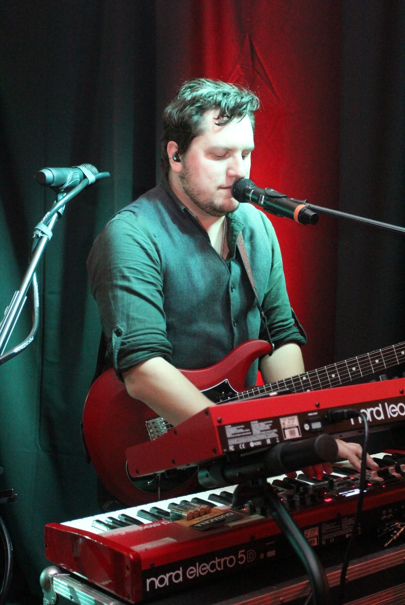 A male musician with dark hair and a beard performs on stage, singing into a microphone while playing a red electric guitar and using a red keyboard. The stage has dark curtains with a red light background.