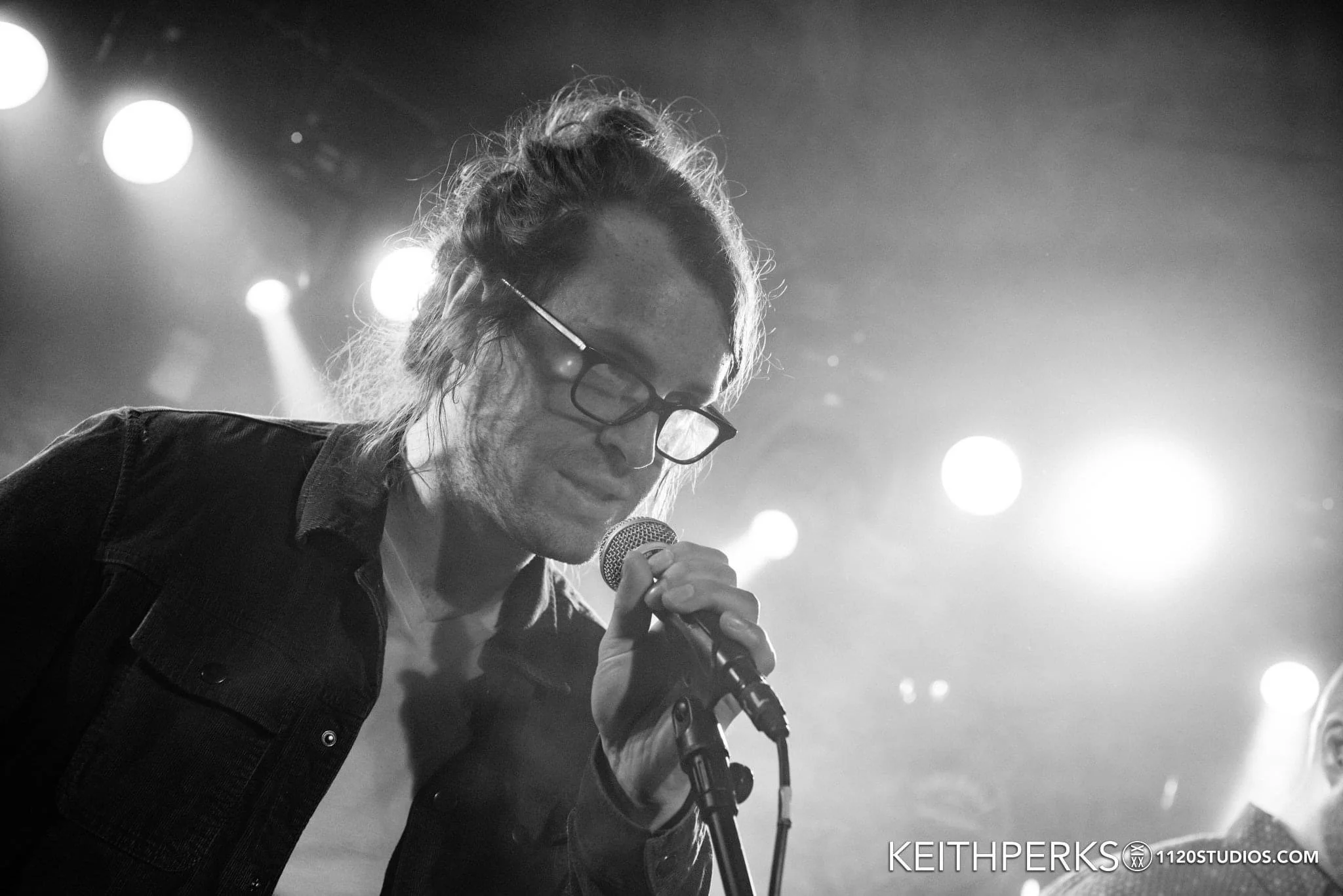 A black and white photo of a man with long hair and glasses singing into a microphone on stage with bright lights behind him.