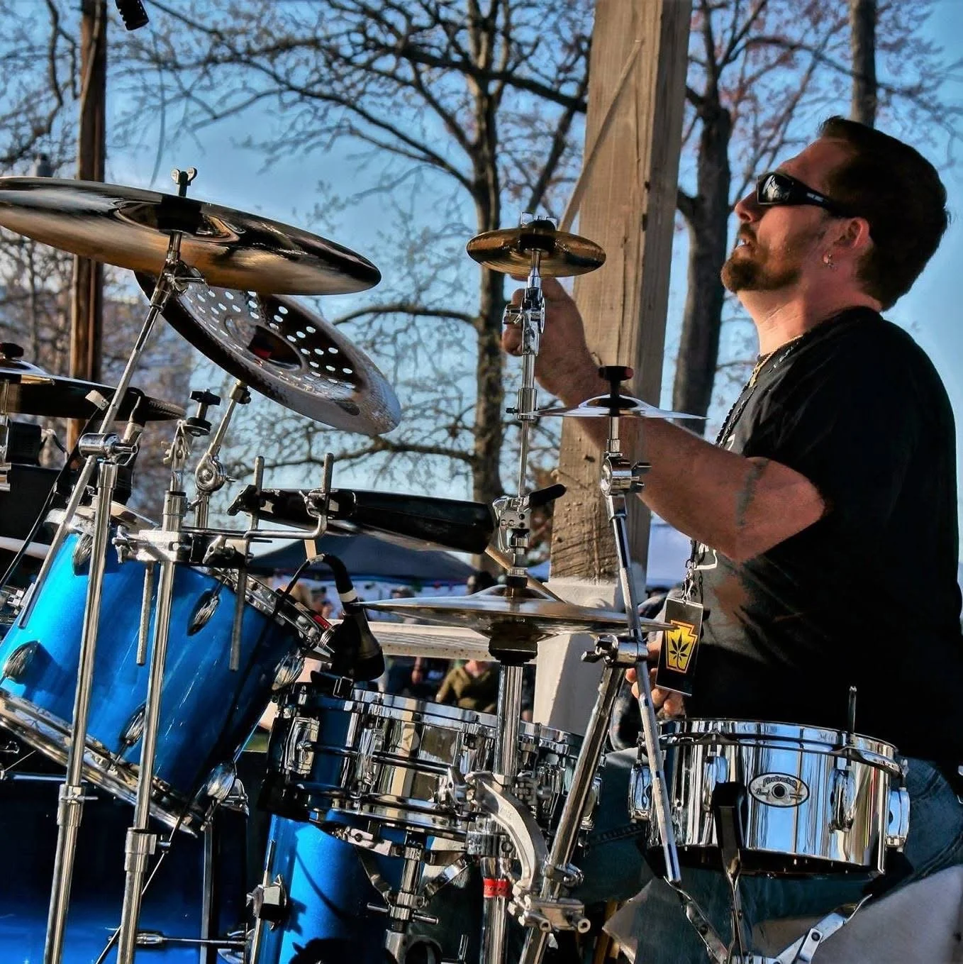 A man with dark hair, goatee, sunglasses, and earrings playing a drum set outdoors during daytime