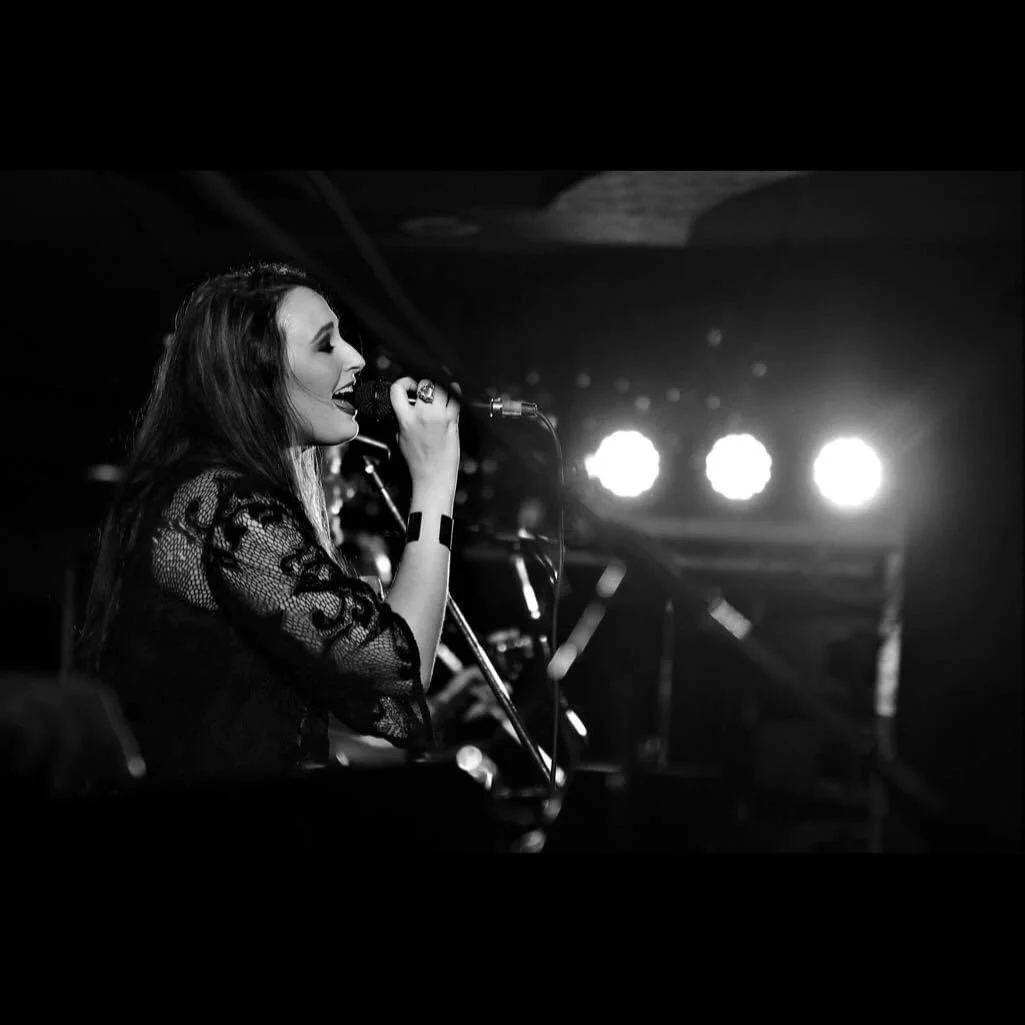 A woman with long hair singing into a microphone on stage, wearing a lace outfit, with bright stage lights behind her in a black and white photo.