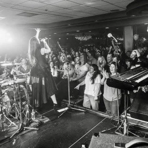 A female singer performs on stage with an engaged audience dancing and singing along, in a concert venue with musical instruments and microphones.