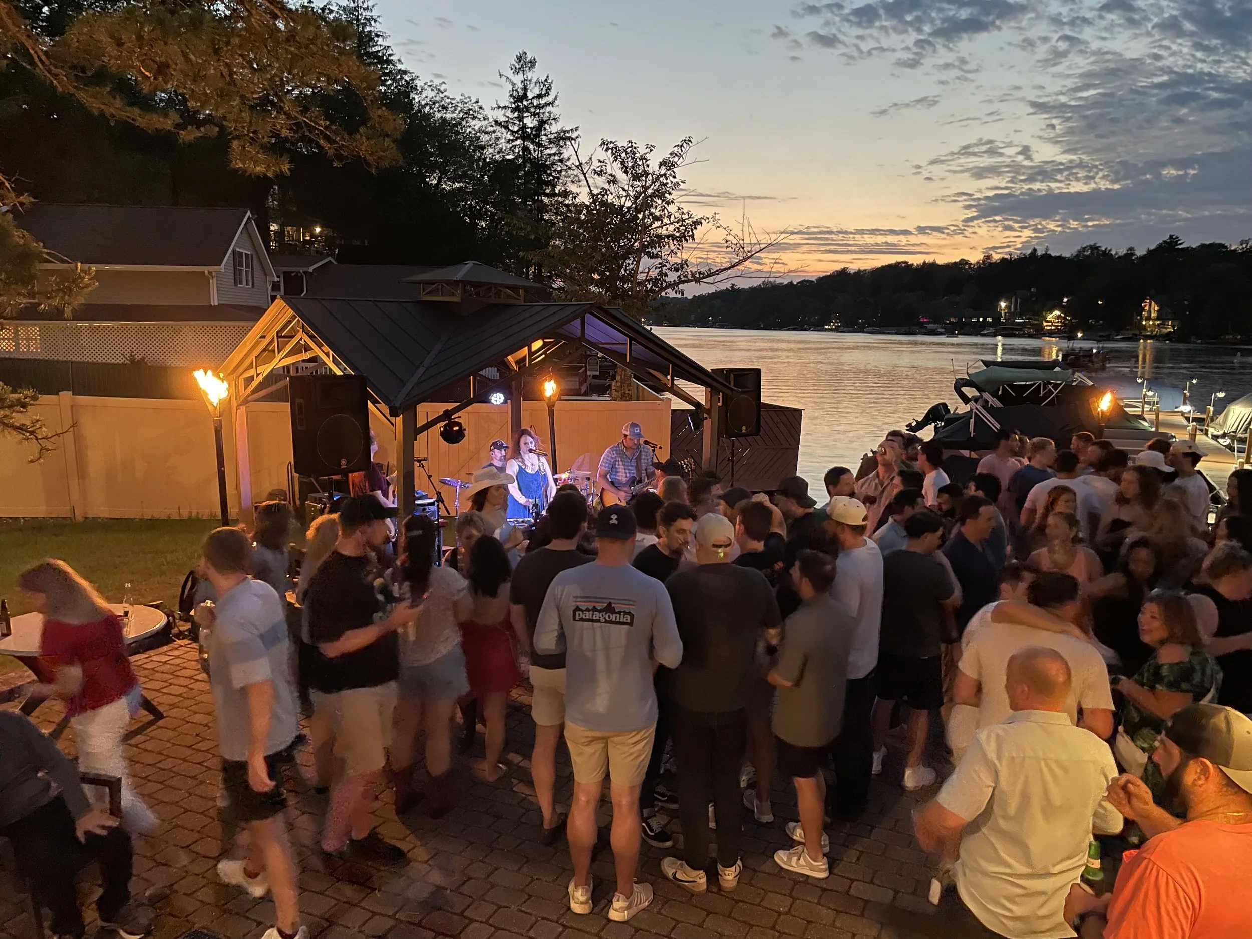 People gathering at an outdoor live music event by a lakeside at sunset, with a stage, musicians, boats, and trees in the background.