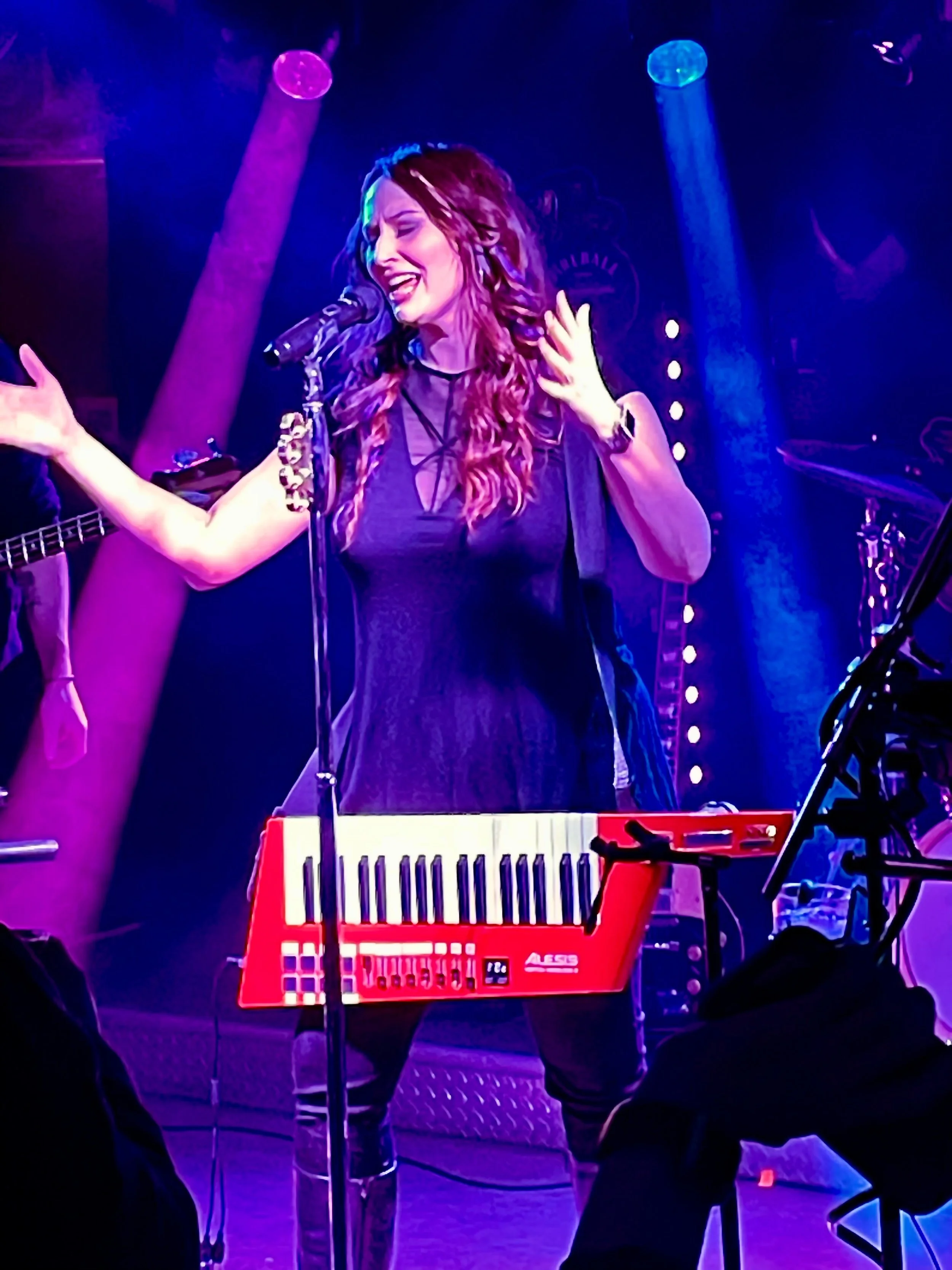 A woman singing passionately on stage with purple and blue stage lights, standing behind a red keyboard with her eyes closed and hand raised.