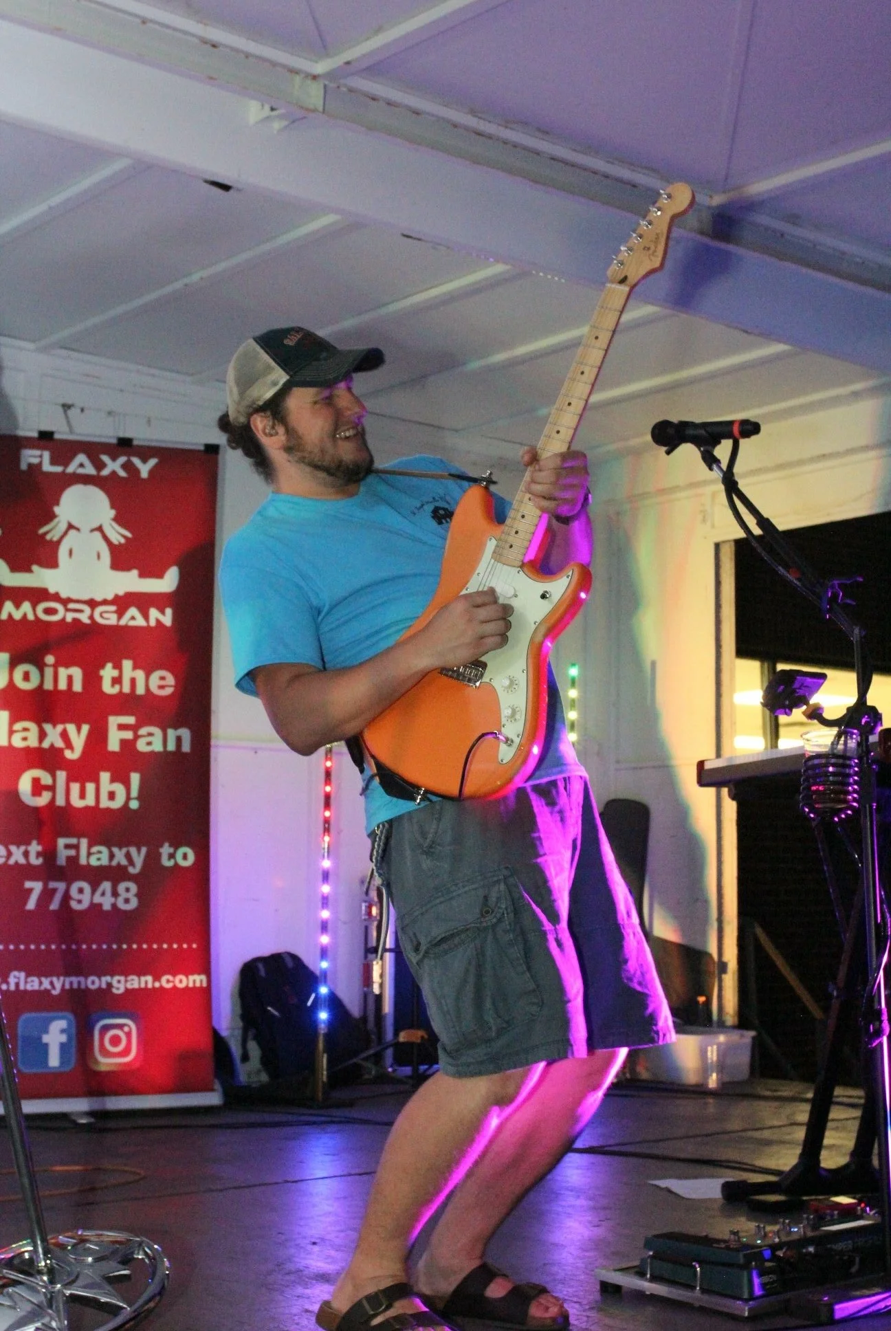 Man wearing a cap and a blue t-shirt, playing an orange electric guitar on stage with colorful lighting at a live music event.