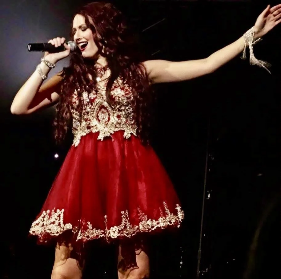 A woman with long, curly red hair singing passionately into a microphone on stage, wearing a red dress with gold embroidery and sheer long sleeves.