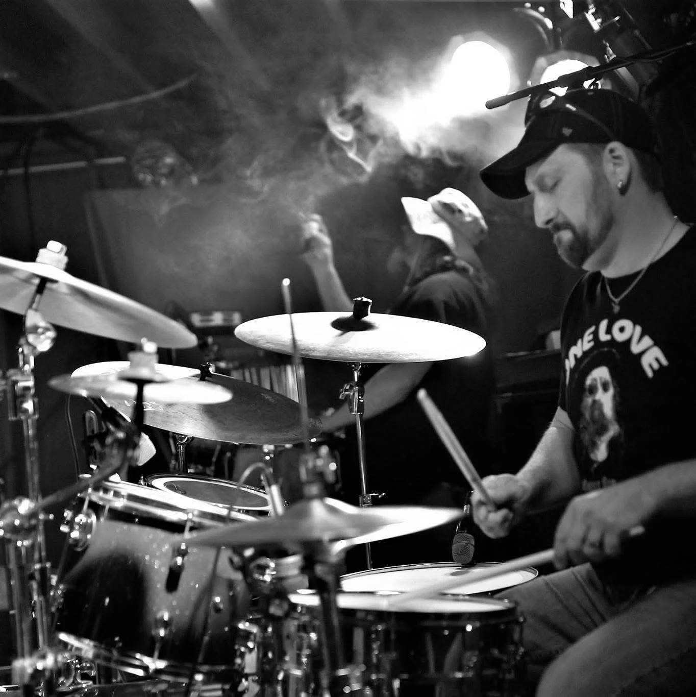 A man playing the drums on stage, wearing a T-shirt with a skull and the words 'Love' and 'Me LoVe'. He is holding drumsticks and appears to be focused. In the background, another person is seen, playing an instrument, with smoke or mist and stage lighting creating a moody atmosphere.