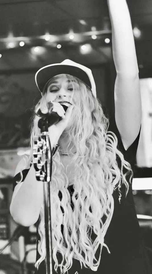 A woman with long curly hair wearing a baseball cap and singing into a microphone on stage, with her arm raised.