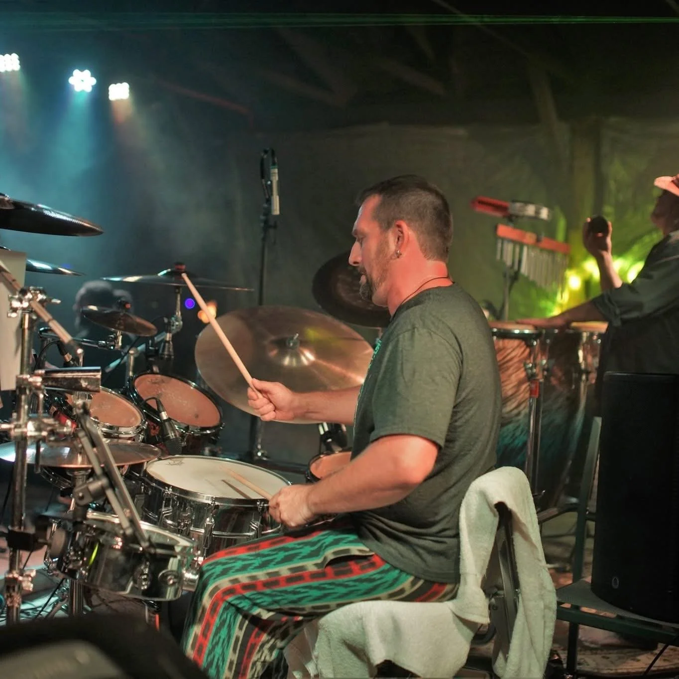A man playing a drum set on stage during a concert, with various musical instruments and equipment visible around him.
