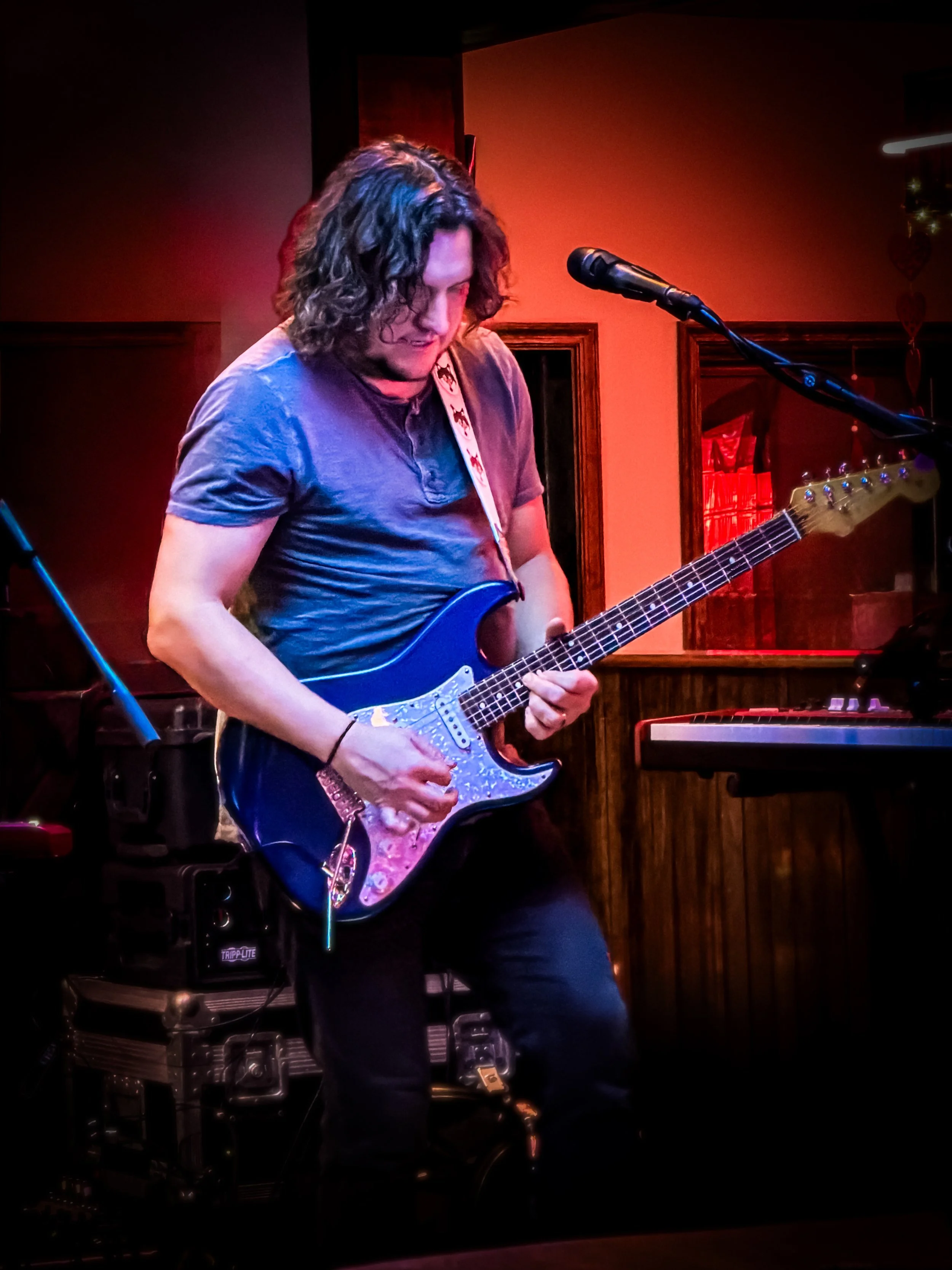 A man with shoulder-length curly hair playing an electric guitar on stage in a dimly lit venue.