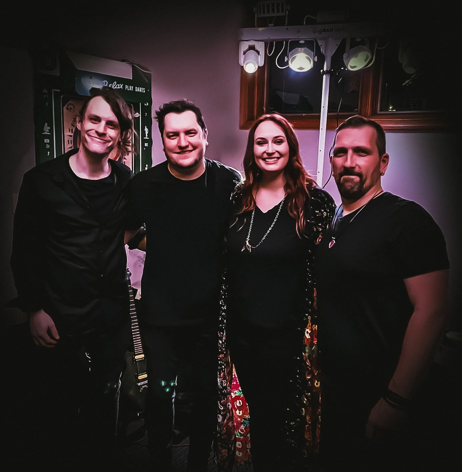 Four people standing together, smiling, with a dartboard visible in the background, under a lighting fixture, in an indoor setting.