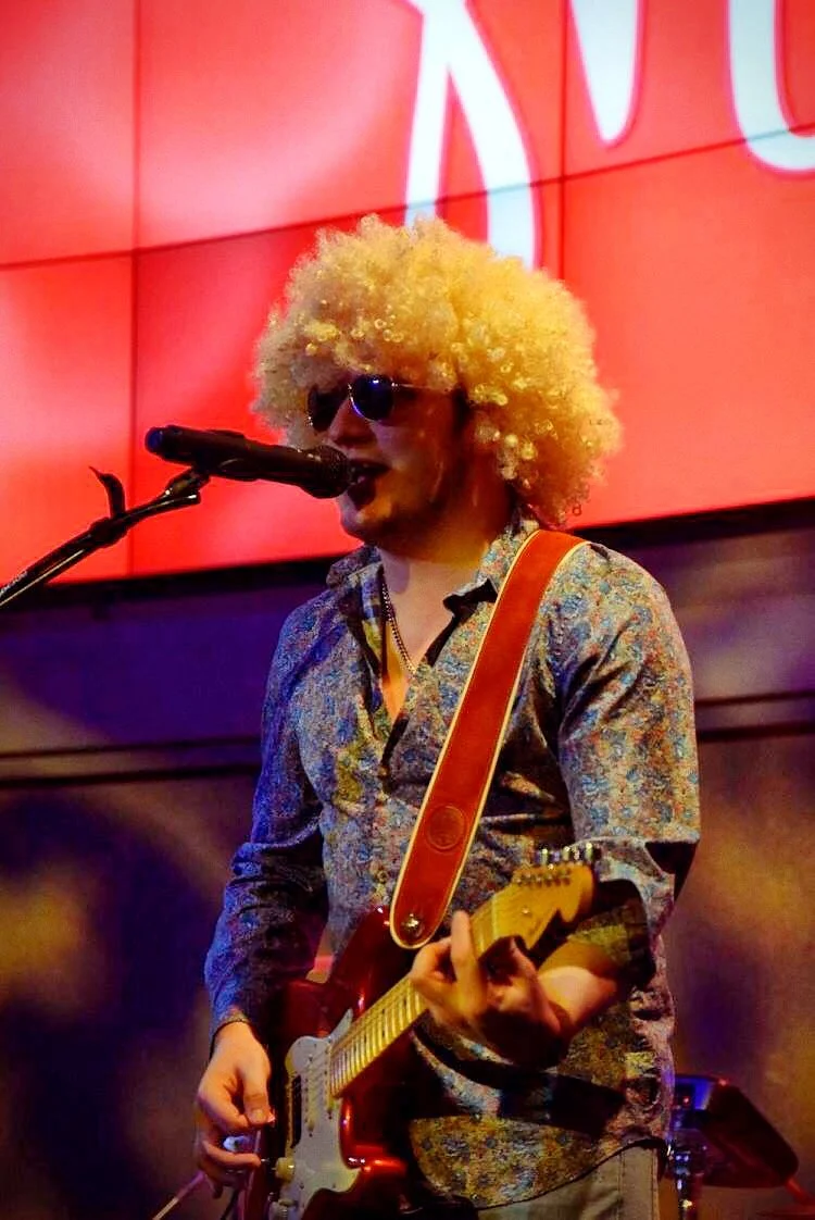 Person with curly blonde wig and sunglasses singing into a microphone, playing an electric guitar, in front of a red background with large white text.