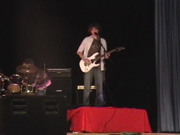 A musician with curly hair and sunglasses playing an electric guitar on stage, with a drummer behind him, a black background, and a red curtain on the side.