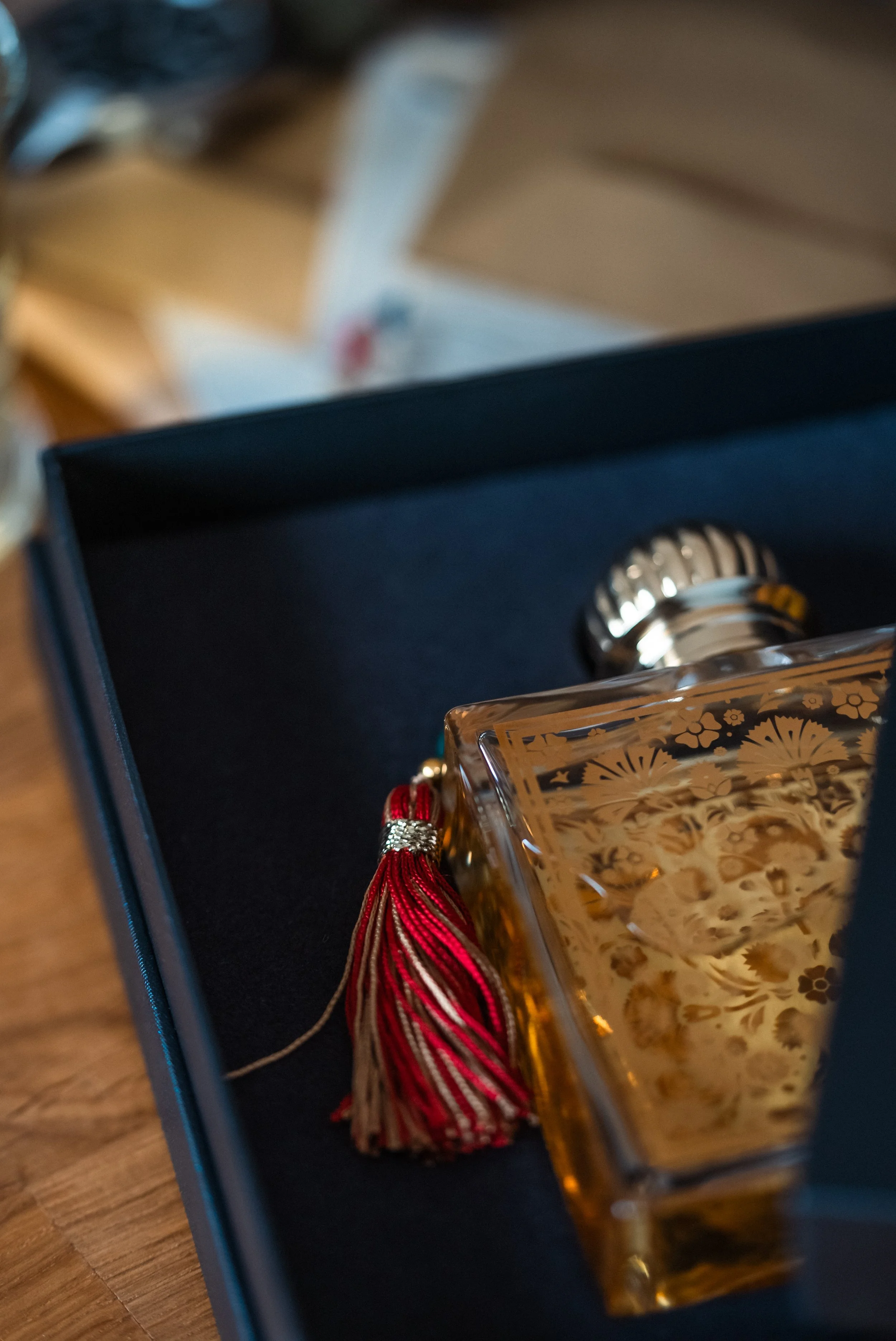 A decorative glass perfume bottle with a gold floral pattern, a shiny silver cap, and a red tasseled ribbon, placed in an open dark box on a wooden surface.