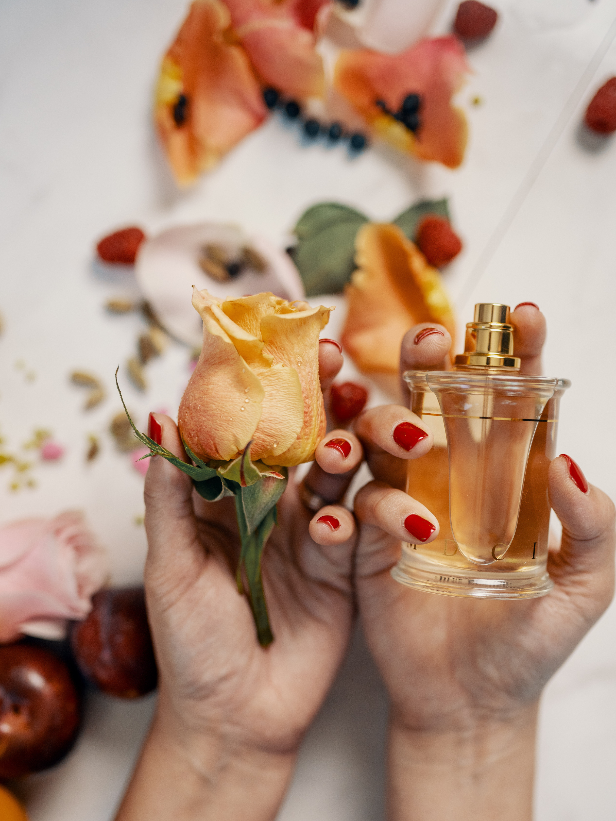 Two hands holding a peach-colored rose and a perfume bottle, with a blurred background of strawberries and other flowers.