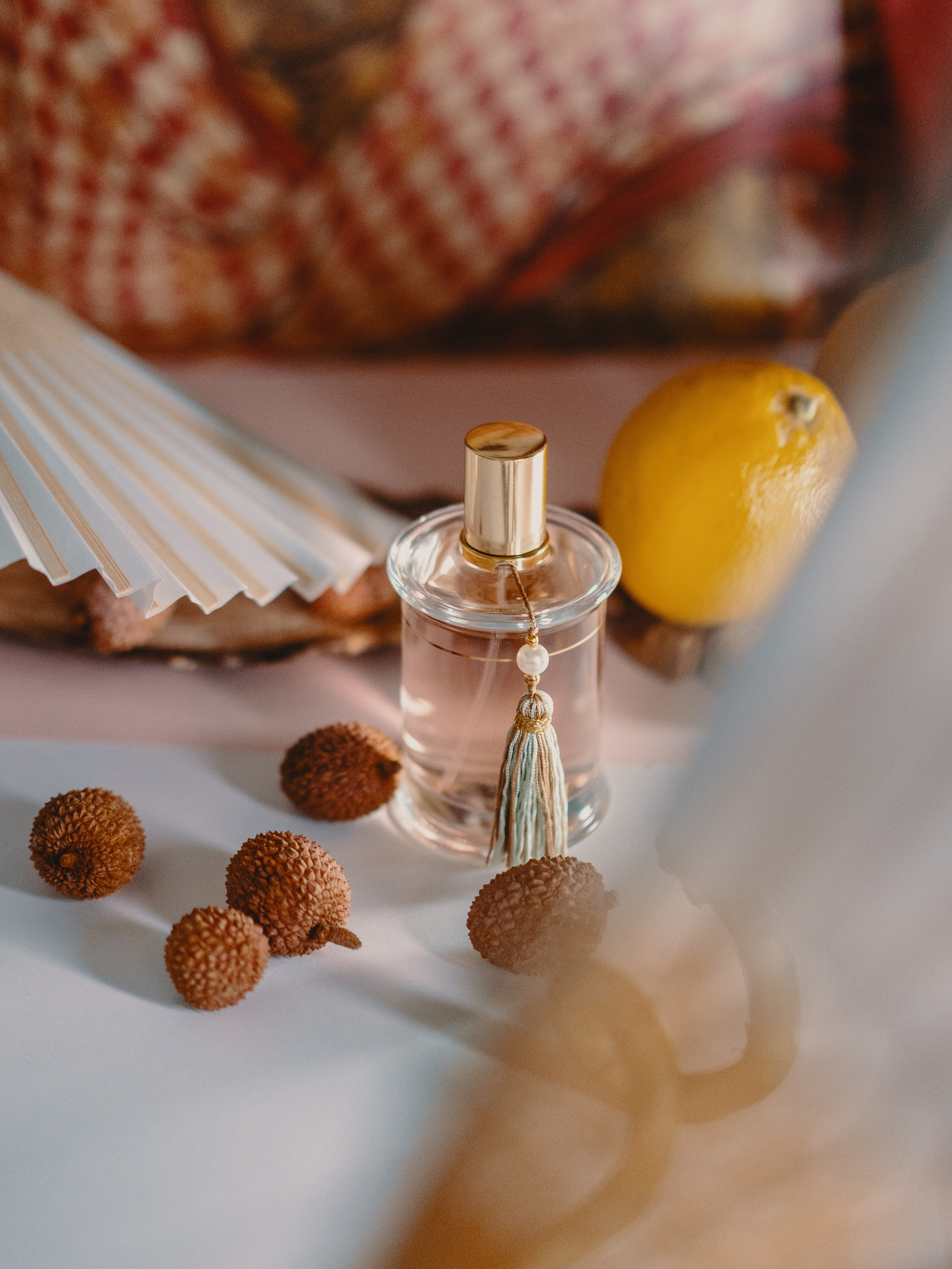 A small clear glass perfume bottle with a gold cap and a tassel hanging from its neck, placed on a white surface. Behind it, there are a lemon and a blurry pink paper with a floral pattern, along with several lychee fruits with brown textured skin scattered on the surface.