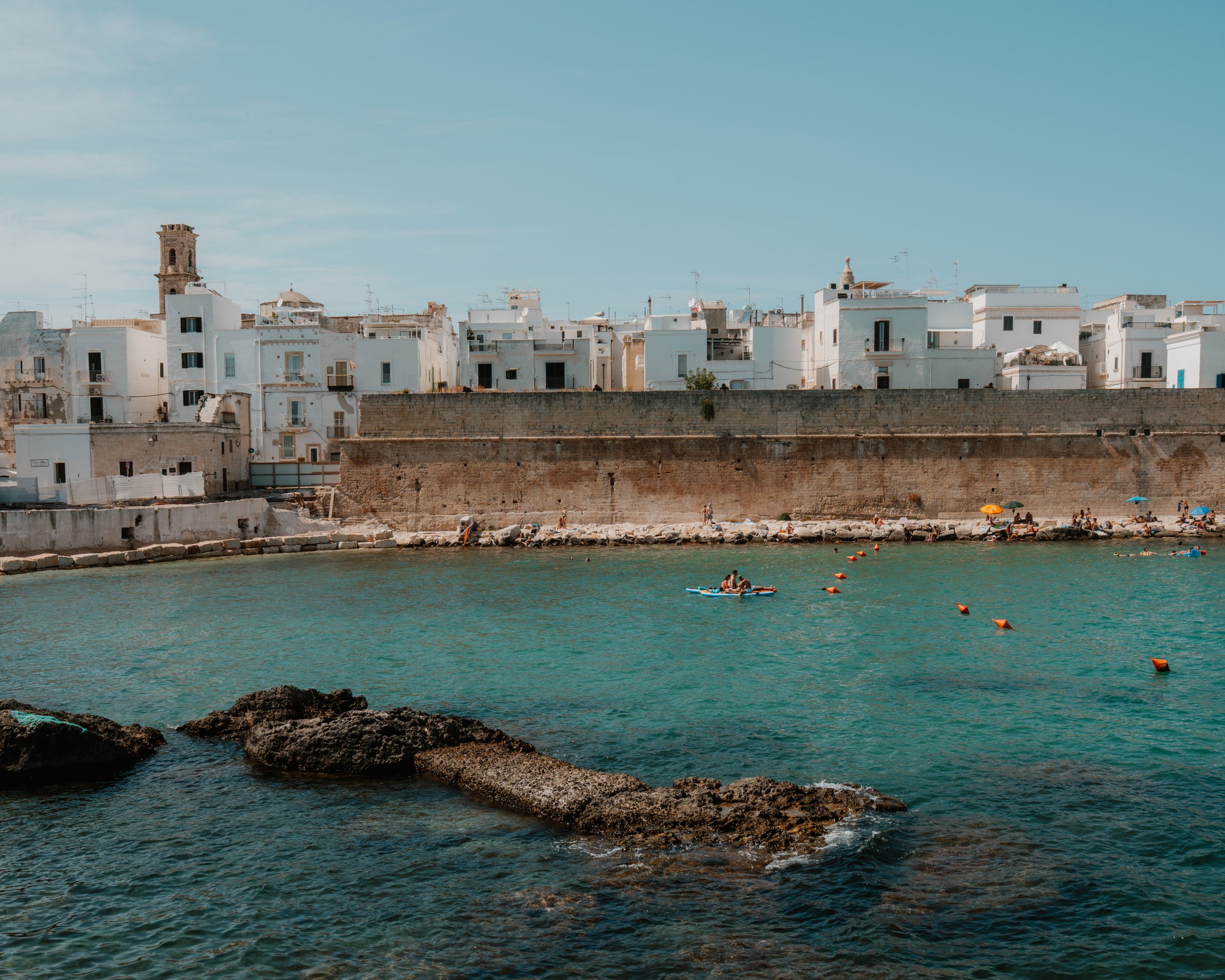 View of a coastal town with white buildings, a stone wall, people on the beach, and calm water with rocks in the foreground, under a clear blue sky.