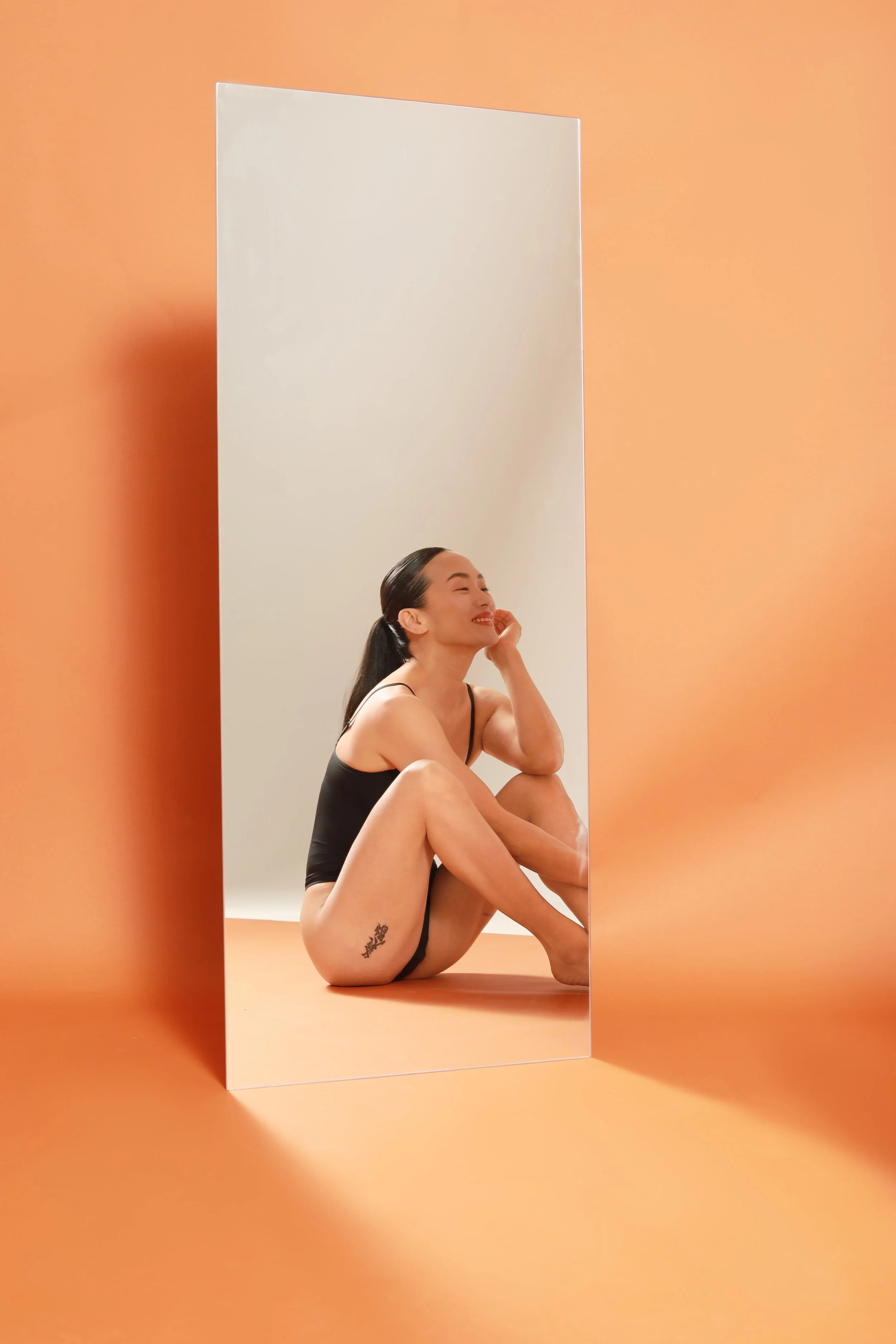 A woman with long dark hair sitting on the floor with her knees bent and arms resting on her knees, seen through a tall mirror against an orange wall. She is smiling, wearing a black camisole, and has a tattoo on her left thigh.